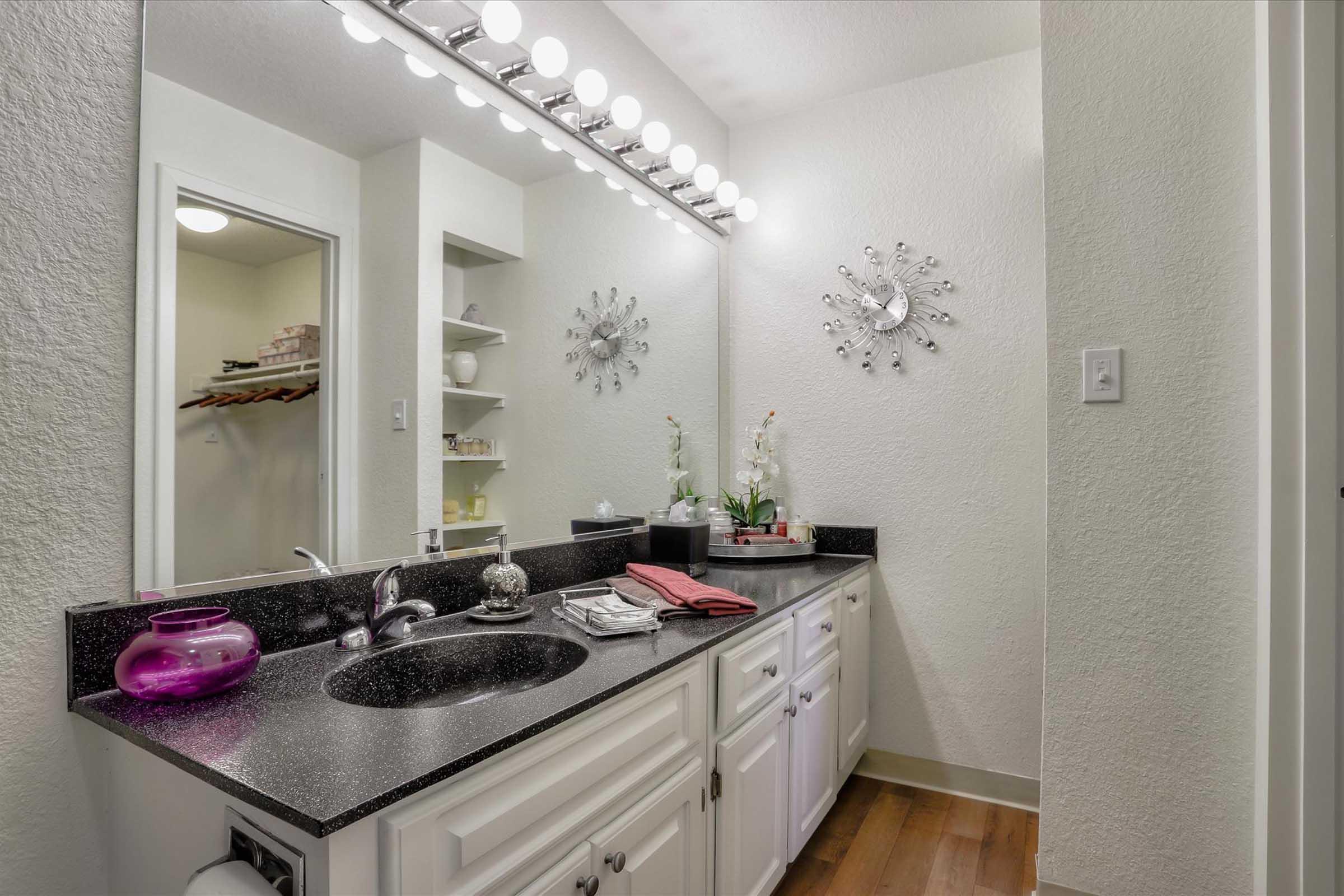 A modern bathroom featuring a dark countertop with a sink, white cabinetry, a pink decorative vase, and neatly arranged toiletries. The wall is adorned with two decorative wall mirrors and bright vanity lighting above the mirror. Light-colored walls and wooden flooring complete the stylish look.
