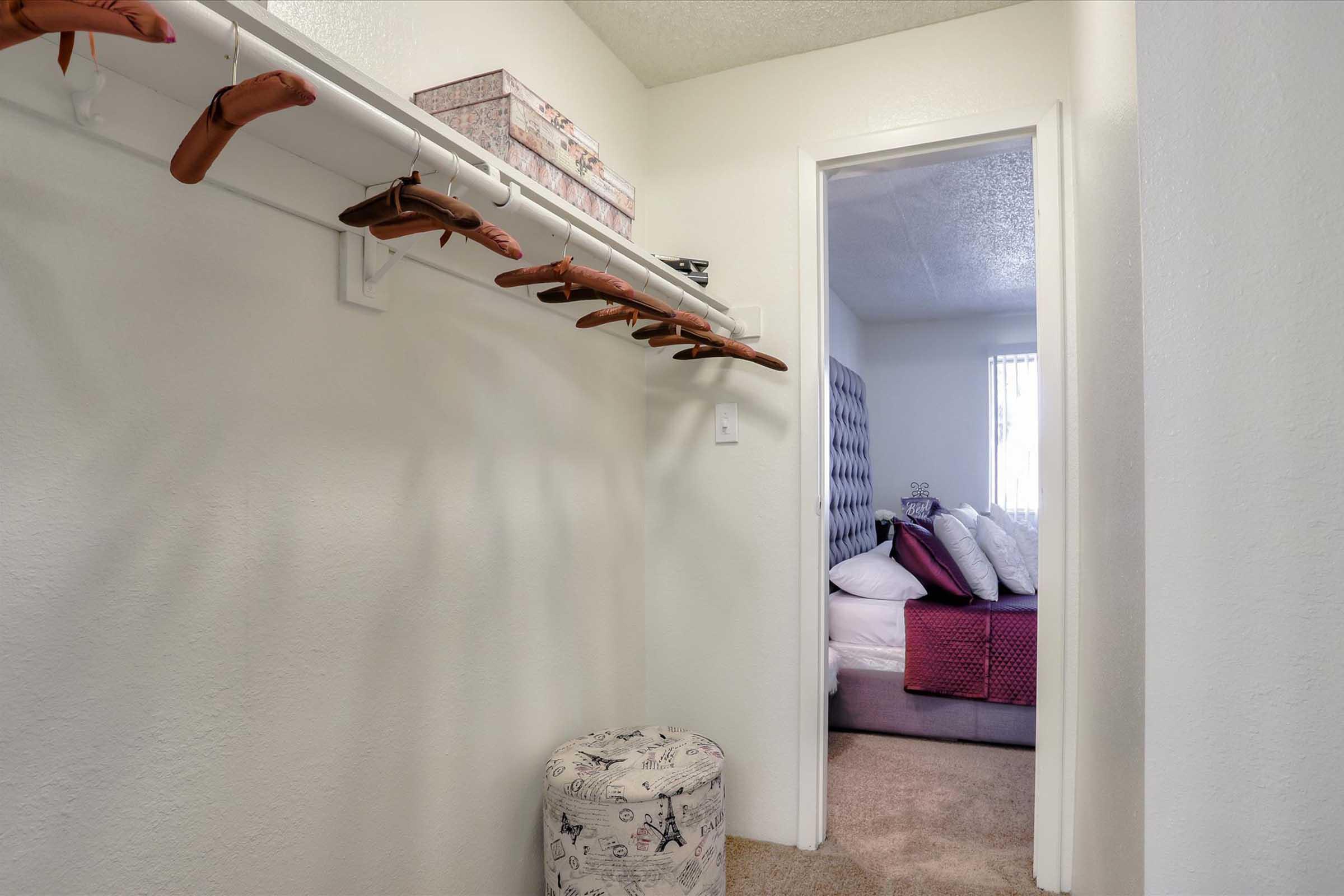 A small hallway featuring a clothes rack with wooden hangers, leading to a bedroom. The bedroom is partially visible, showing a bed with patterned bedding and several pillows. There is also a round ottoman with a butterfly design in the hallway, and soft lighting throughout the space.