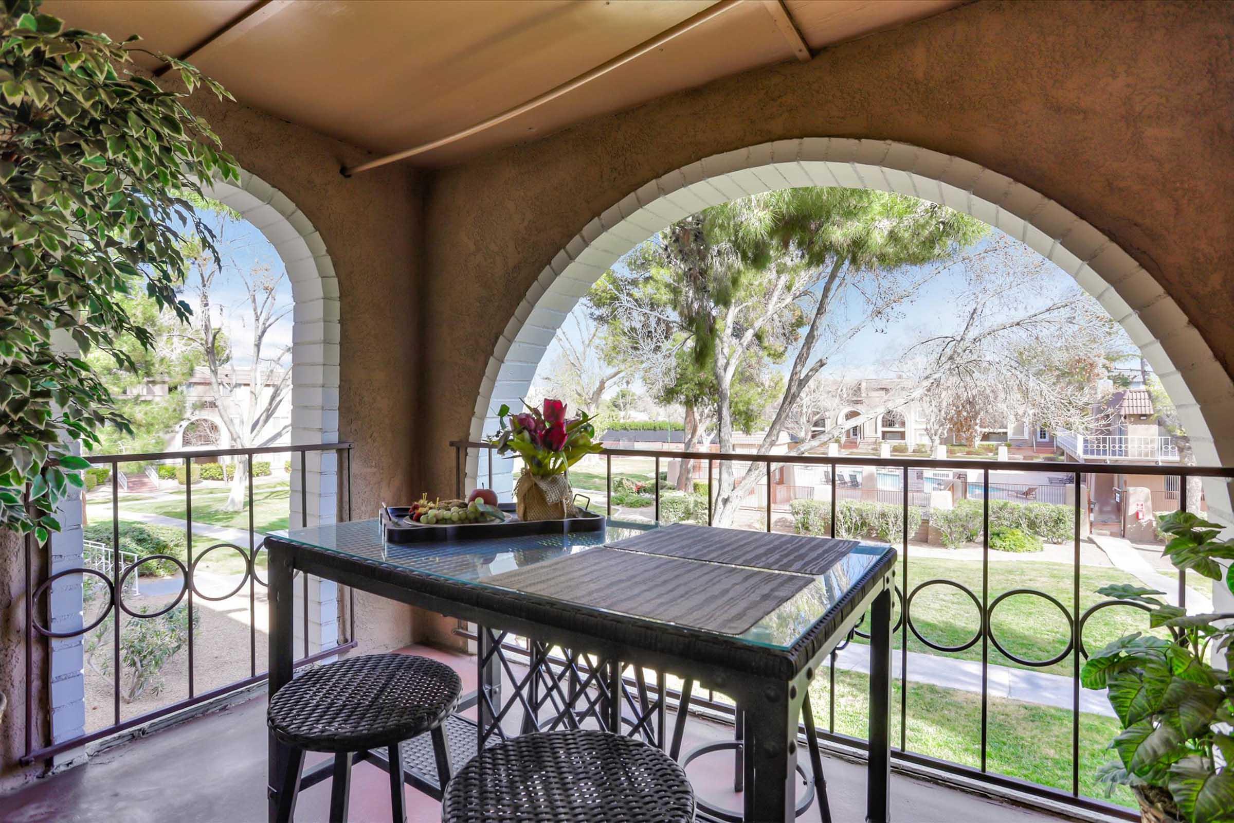 A cozy balcony with arched openings overlooking a landscaped area. A glass-top table with two black chairs is set, featuring a vase of flowers. Lush greenery adorns the balcony, creating a tranquil spot to relax and enjoy the view of the surroundings.