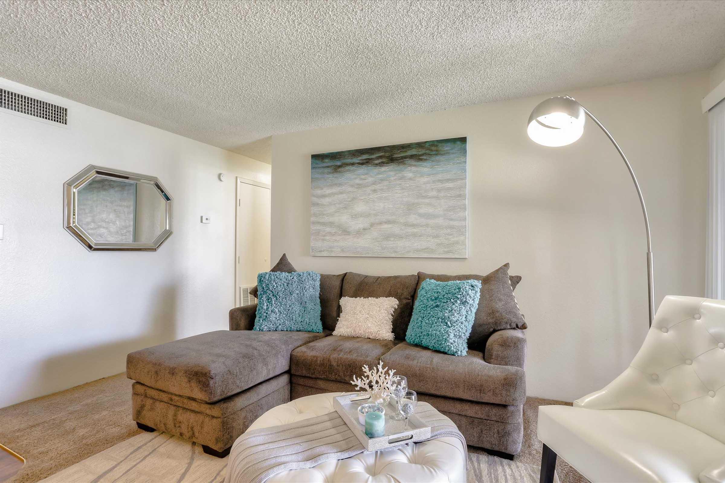 A cozy living room featuring a brown sectional sofa with turquoise and gray throw pillows. There's a white decorative ottoman in the foreground, and a modern floor lamp beside the couch. A large abstract painting hangs on the wall, and a mirror reflects light. The room has neutral-colored walls and carpet.