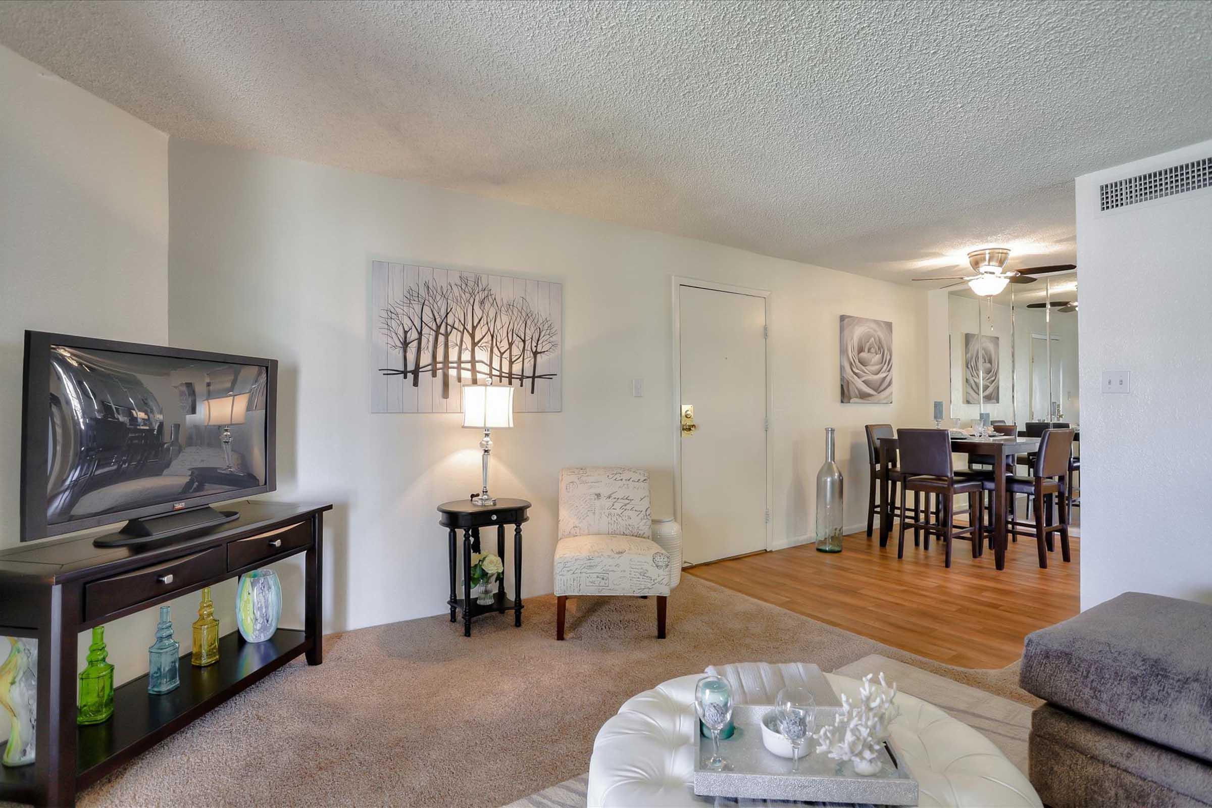 A cozy living room featuring a television, decorative lamp, and a chair with a floral design. In the background, there's a dining area with a table and chairs, along with artwork on the walls. The space has a warm, inviting atmosphere with wood flooring and pleasant decor elements.