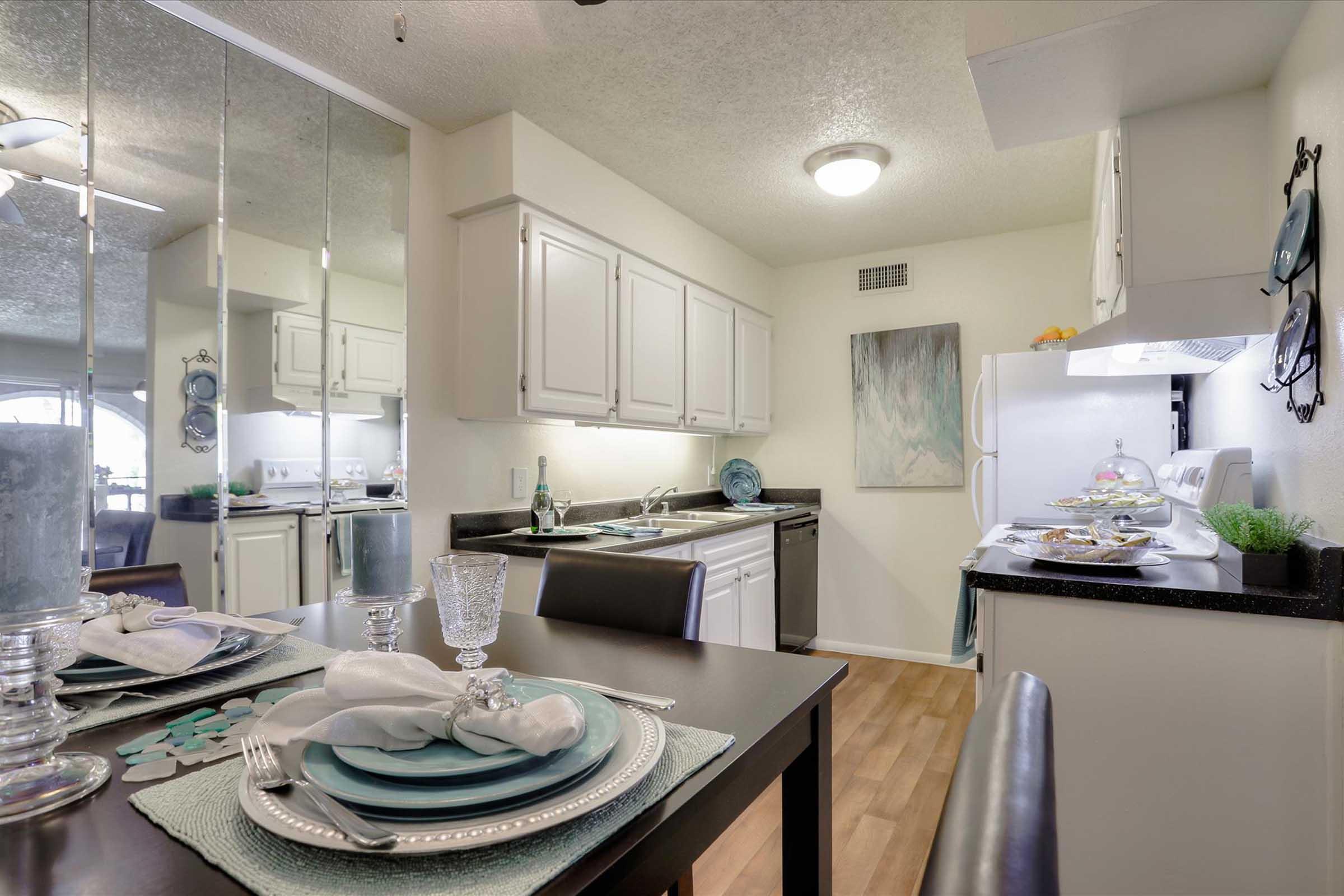 A well-lit kitchen and dining area featuring a black table set for a meal. The table is adorned with elegant dishware and glassware. The kitchen has white cabinetry and modern appliances, with a reflective wall enhancing the space. Soft neutral colors create a cozy ambiance.