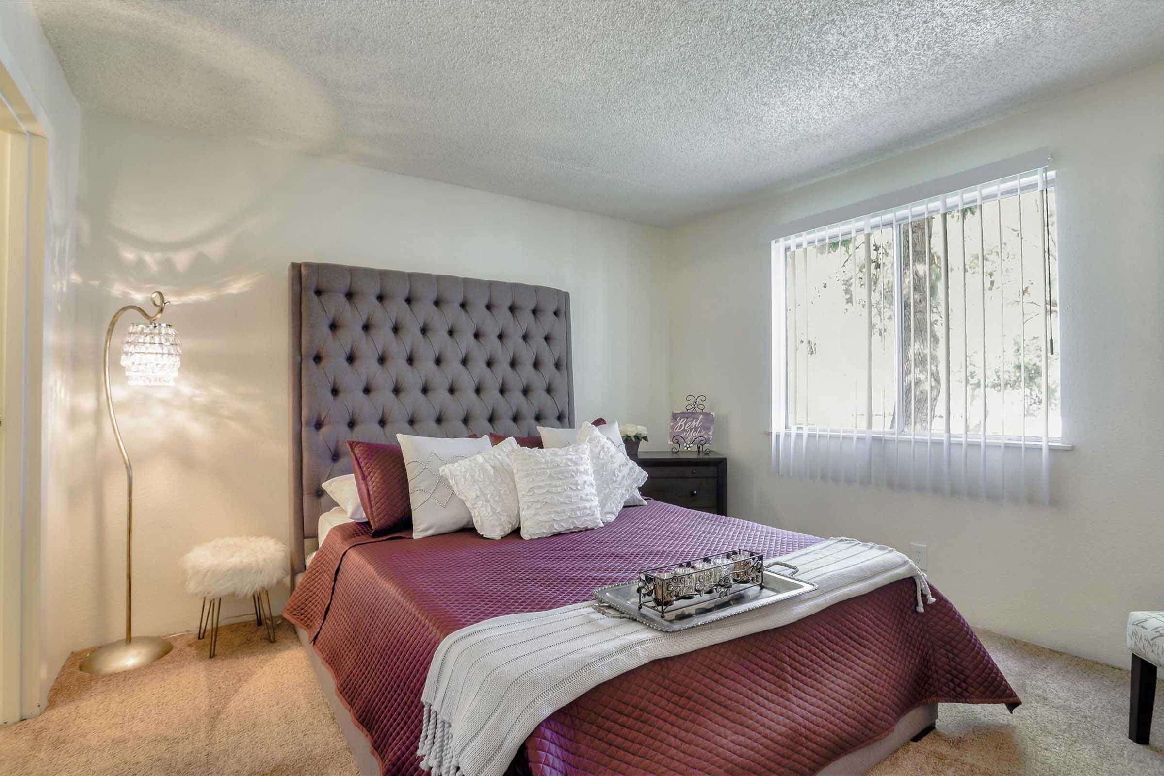 Cozy bedroom featuring a large tufted headboard, a neatly made bed with decorative pillows, and soft purple bedding. A bedside table with a decorative tray sits next to the bed, and a stylish floor lamp adds ambient light. A window with white blinds allows natural light to filter in.