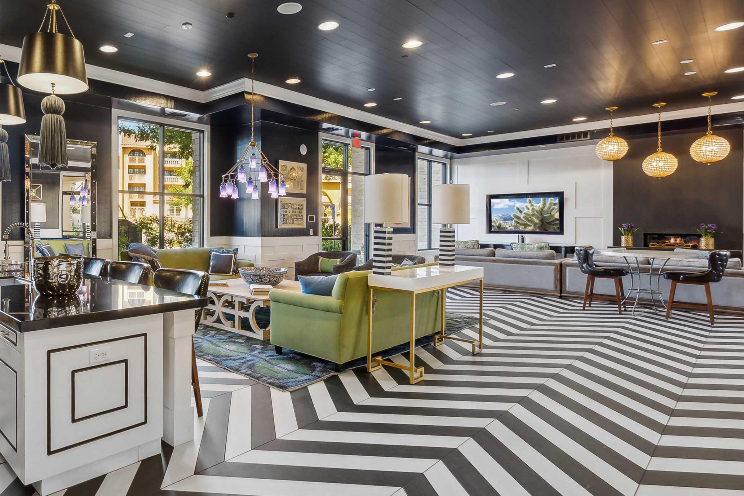 A modern lounge area with a stylish black and white chevron floor. The space features comfortable seating in green and gray, large windows letting in natural light, decorative lighting, and a flat-screen TV on the wall.