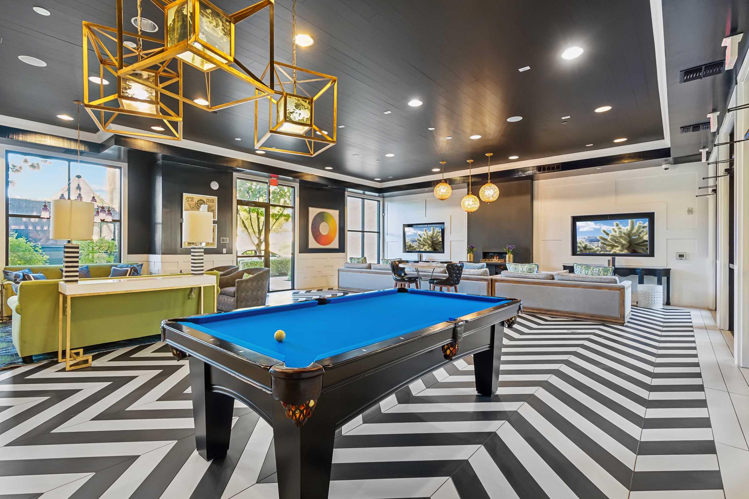 A modern lounge area featuring a blue pool table in the center, surrounded by stylish seating, decorative lighting fixtures, and large windows with natural light.