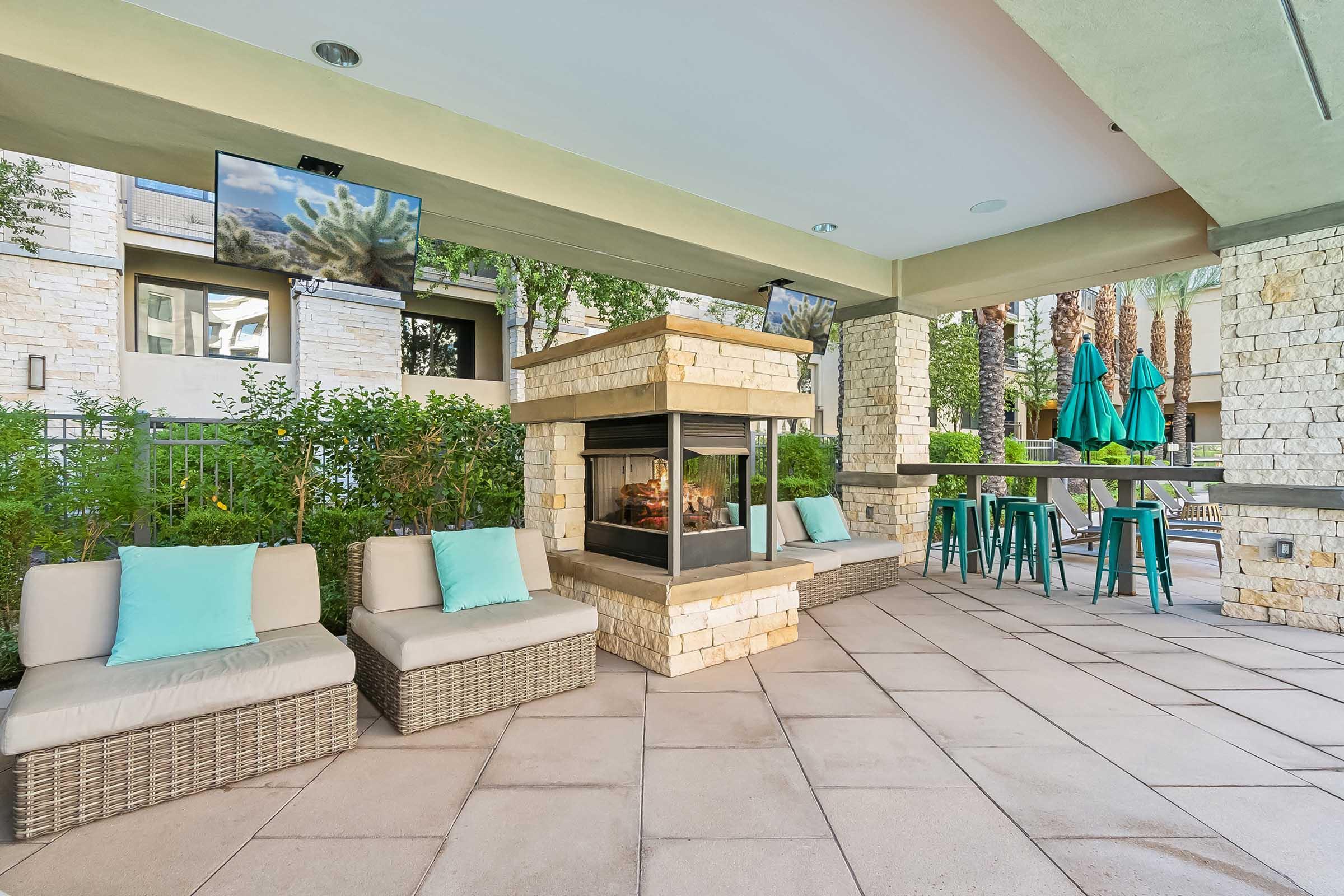 An outdoor patio area featuring a stone fireplace, cushioned seating with light blue accents, and bar stools.