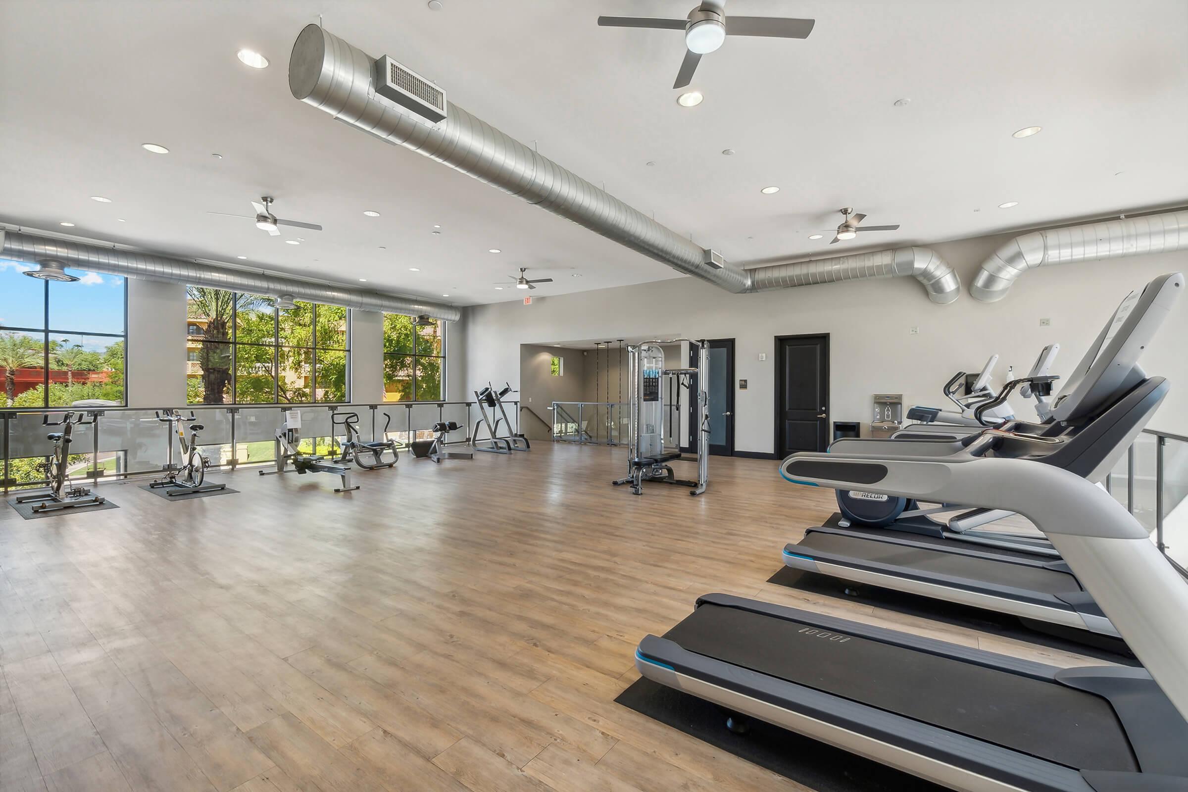 Modern fitness center with wooden flooring and large windows letting in natural light. The gym is equipped with various exercise machines, including treadmills and elliptical trainers, along with weights and other fitness equipment.