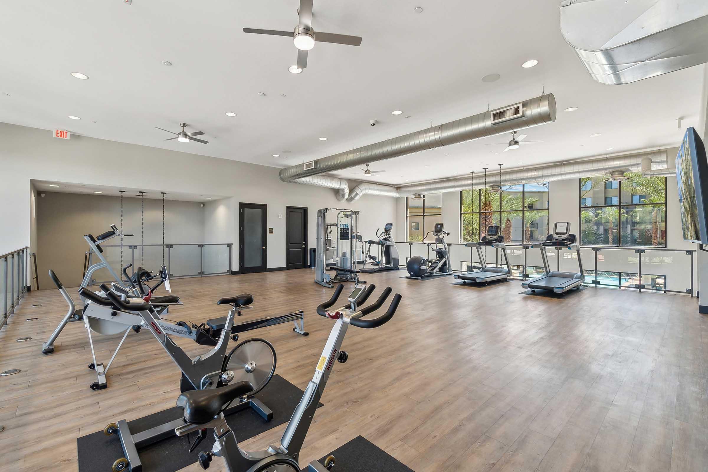 A bright, modern gym interior featuring various exercise equipment including stationary bikes, treadmills, and elliptical machines.