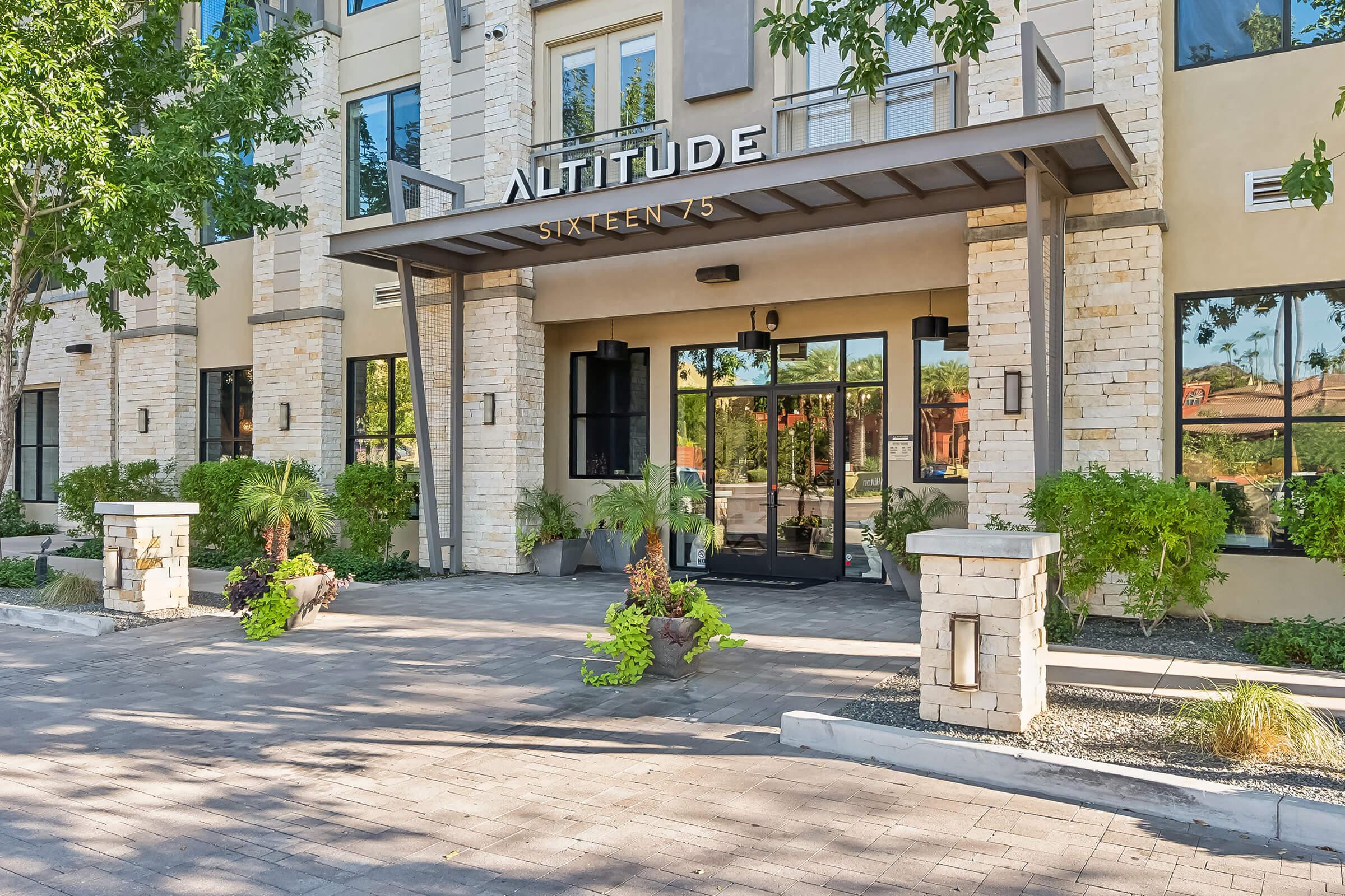 Modern building entrance with a sign reading "Altitude Sixteen 75". The façade features stone accents and large glass doors.