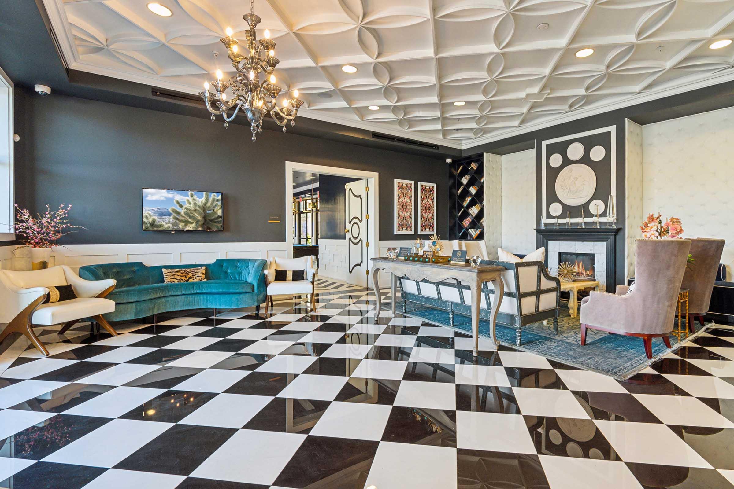 A stylish interior room featuring a checkered black and white floor, elegant furniture in a mix of colors including teal and beige, a chandelier overhead, and framed artwork on the walls.