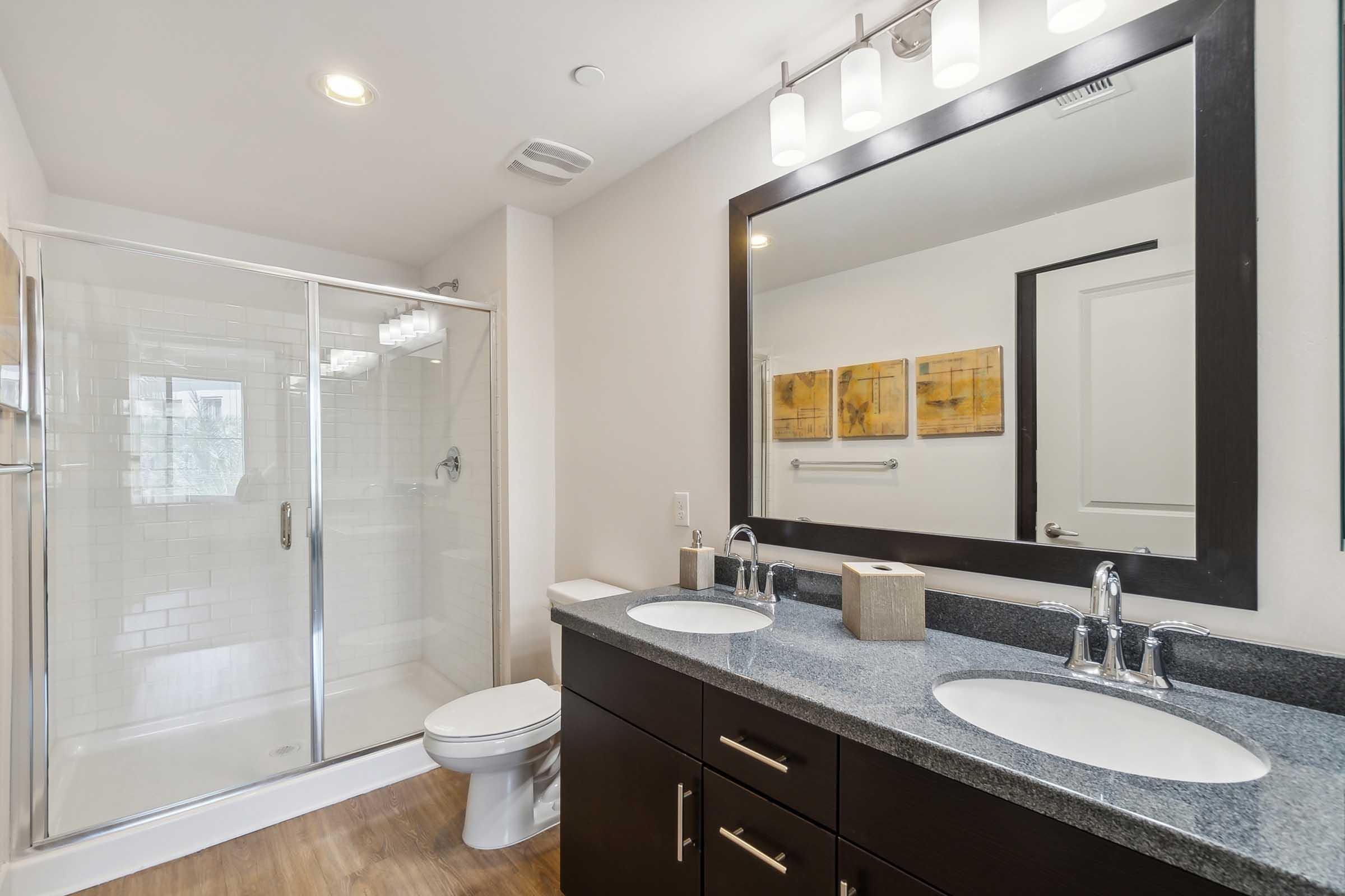 Modern bathroom featuring a double sink vanity with dark cabinets and a gray countertop, a large mirror above, and a glass-enclosed shower.