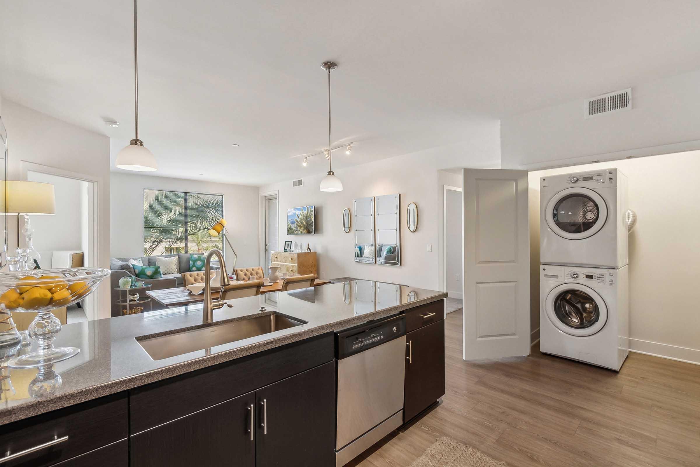 Modern apartment interior featuring a kitchen with a dark countertop, stainless steel appliances including a dishwasher, and an adjoining laundry area with stacked washer and dryer.