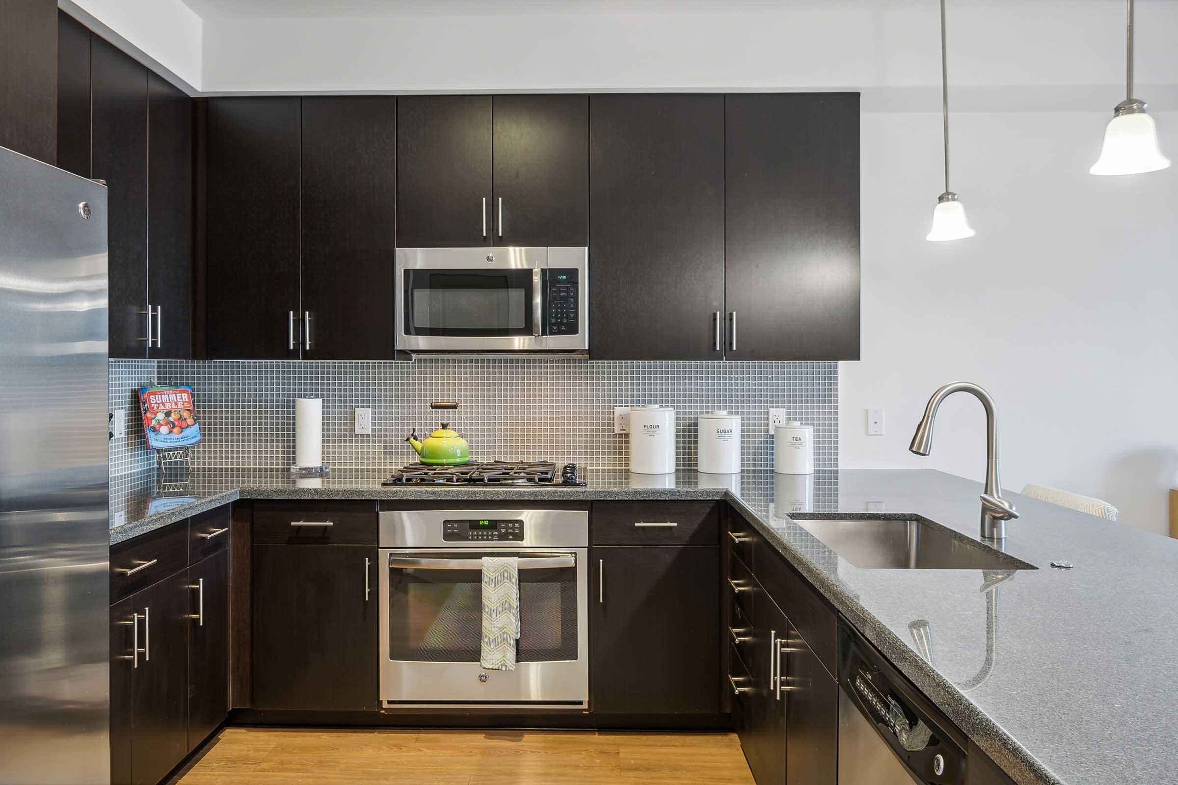 A modern kitchen featuring dark wood cabinets, stainless steel appliances, a subway tile backsplash, and a large granite countertop. The