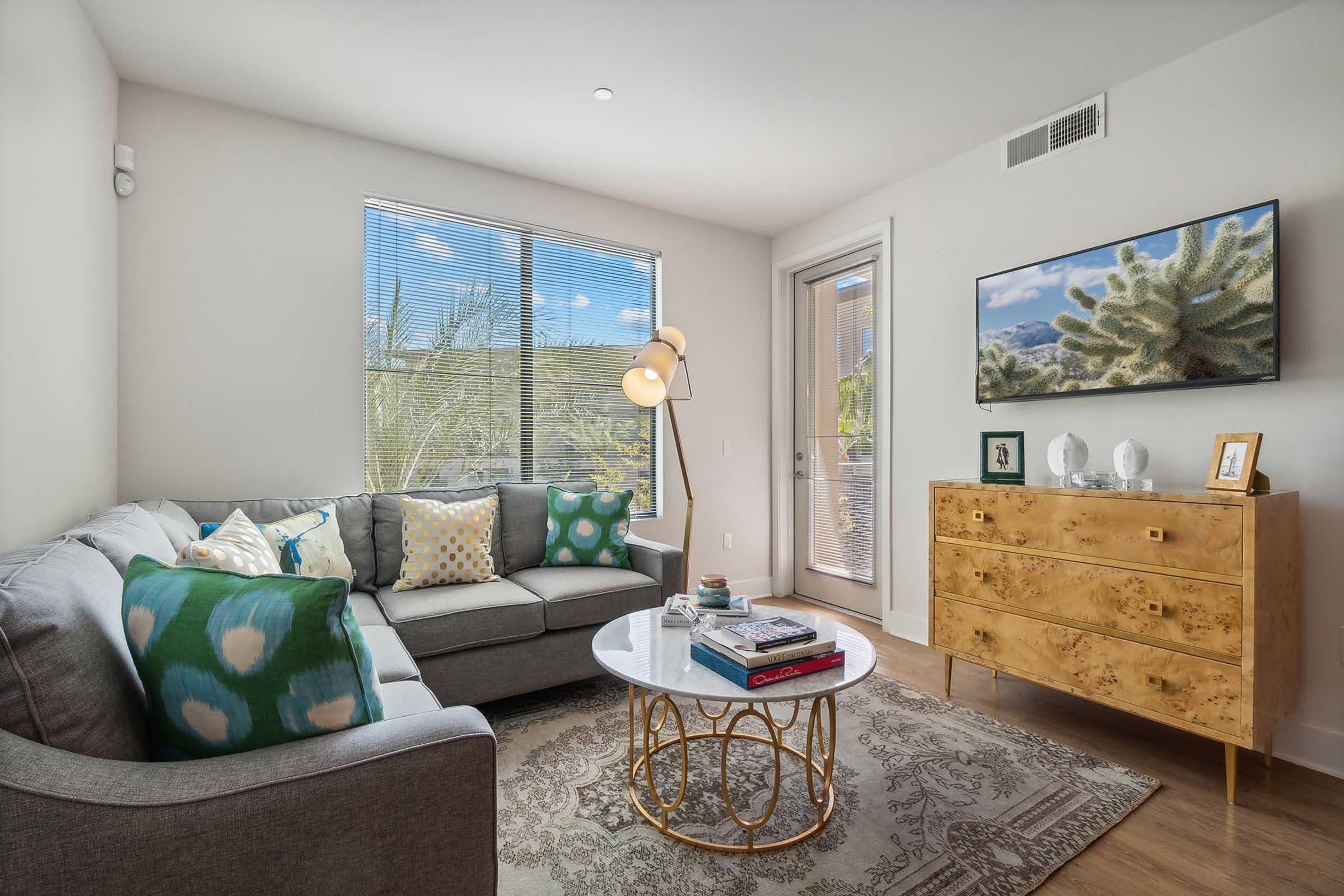 Cozy living room featuring a light gray sofa adorned with colorful cushions, a round glass coffee table with books, and a decorative wooden chest of drawers.