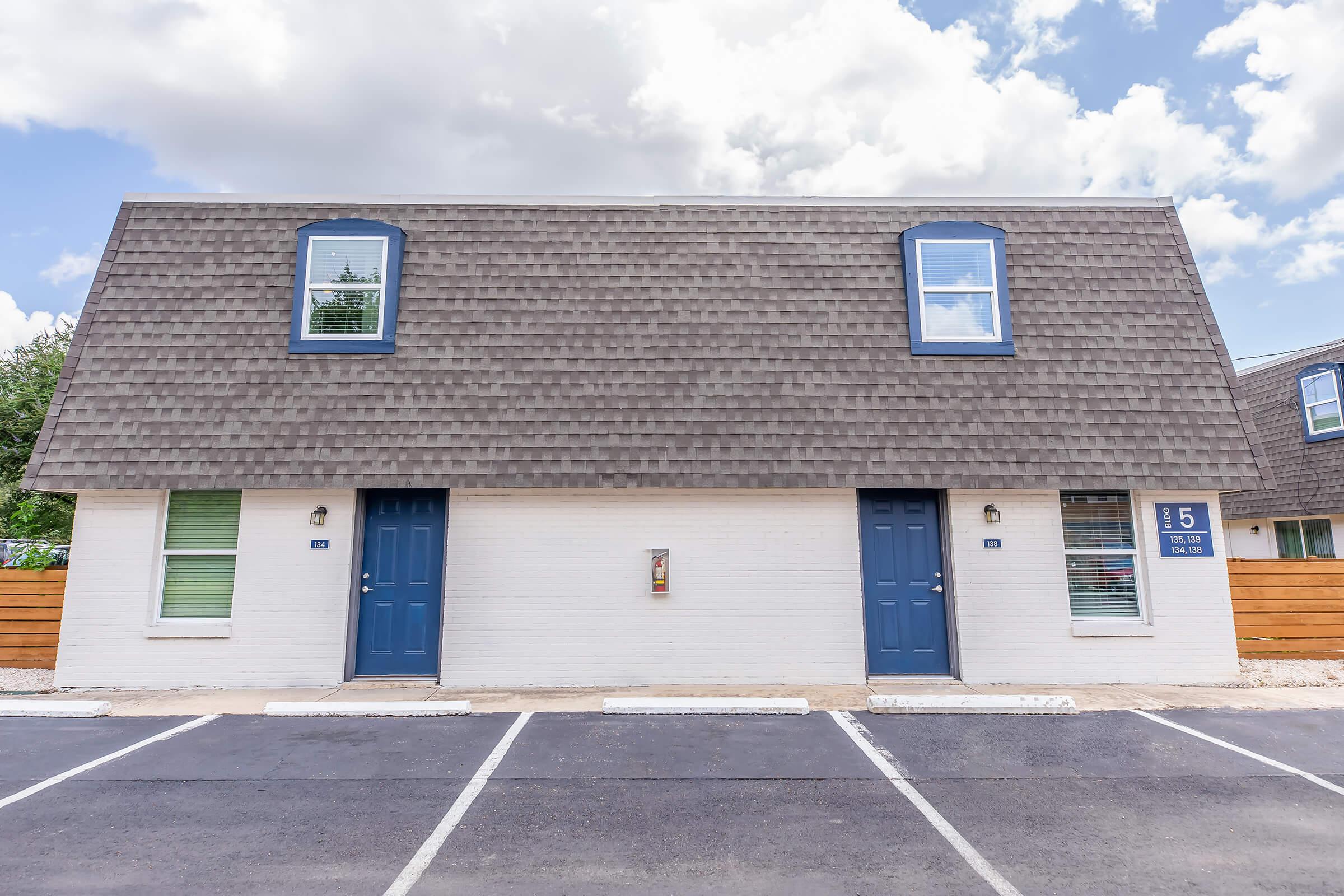 Two-story apartment building with a pitched roof, featuring two identical units, each with blue doors and windows. The exterior is painted white, and there are parking spaces in front. The building is set against a partly cloudy sky, with a wooden fence visible to the side.