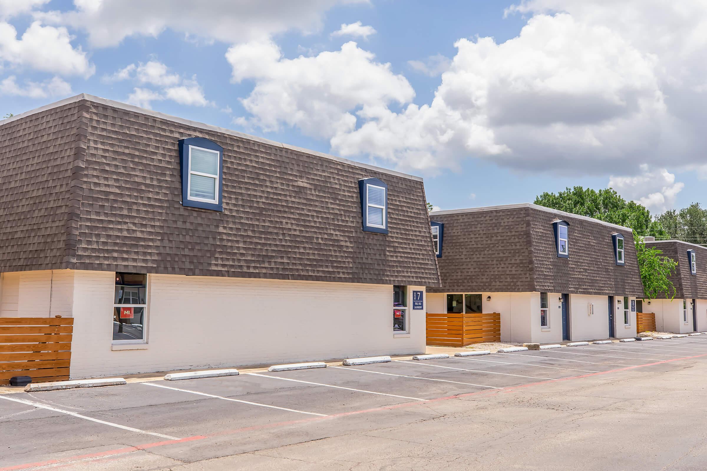 A row of modern, low-rise buildings with distinctive sloped roofs, featuring multiple windows. The buildings are painted in light colors, with some wooden accents. A parking lot is in front, with marked spaces, and the sky is partly cloudy with a few trees in the background.