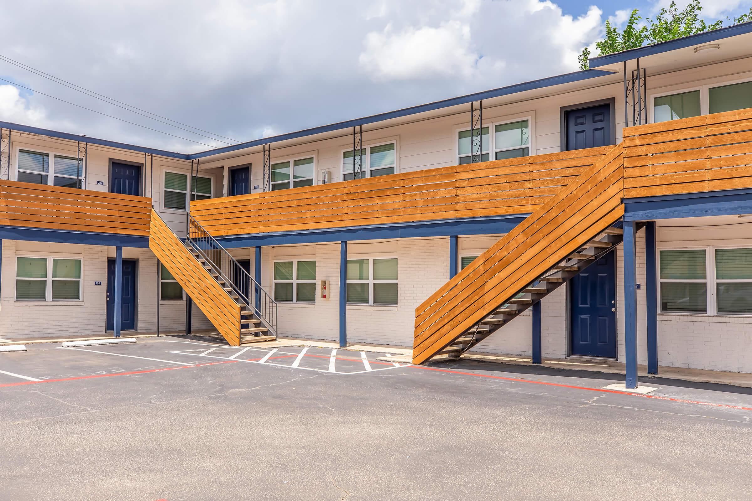 Two-story motel with tan and blue exterior, featuring wooden staircases leading to upper-level rooms. The parking area is visible in front, with marked spaces. The sky is partly cloudy, adding a bright ambiance to the scene.