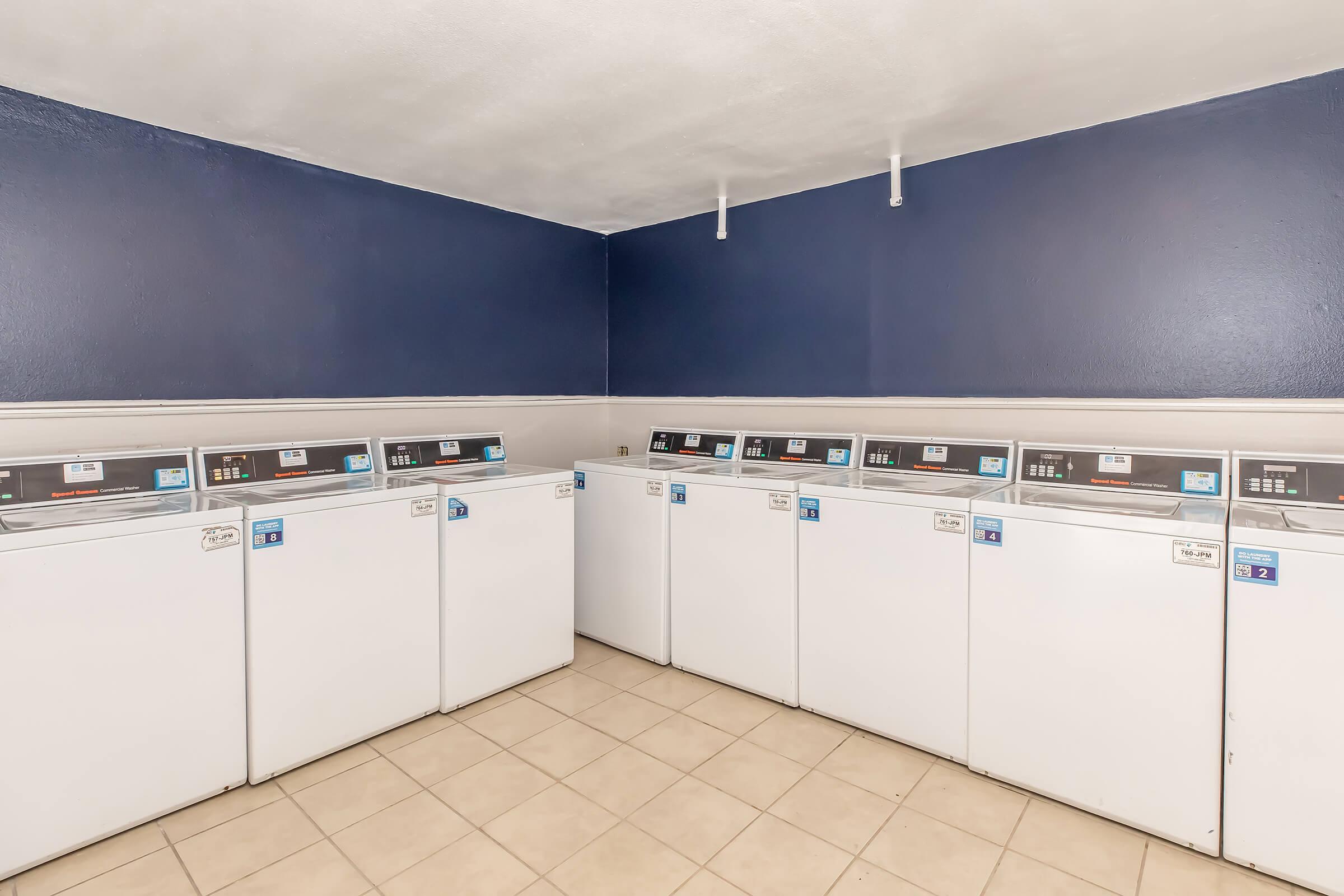 A clean laundry room featuring several white washing machines lined up against a dark blue wall. The floor is tiled, and the overall environment appears well-maintained and organized, suitable for doing laundry.
