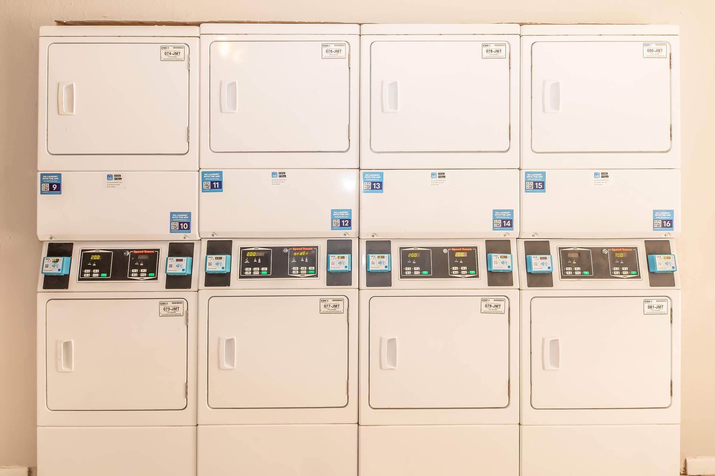 A row of stacked white dryers with digital control panels, displaying various settings and time indicators. Each unit features a clear label with pricing information and operational status lights. The dryers are arranged neatly against a light-colored wall.