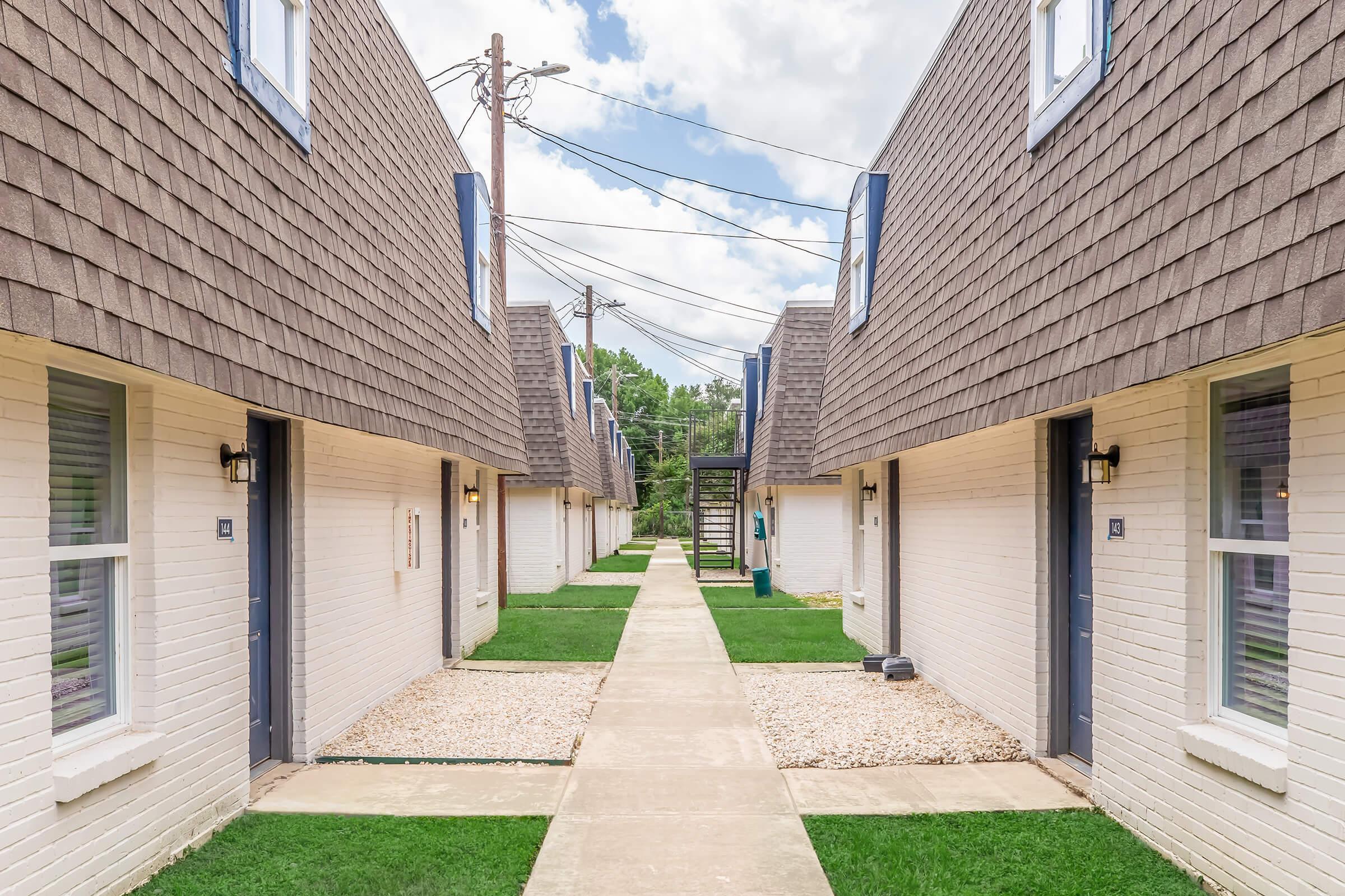 A view of a residential courtyard featuring two rows of buildings with sloped roofs. The path between the buildings is lined with grass and stones, and there are power lines in the background under a partly cloudy sky. The buildings have light-colored walls and dark blue accents around the windows.