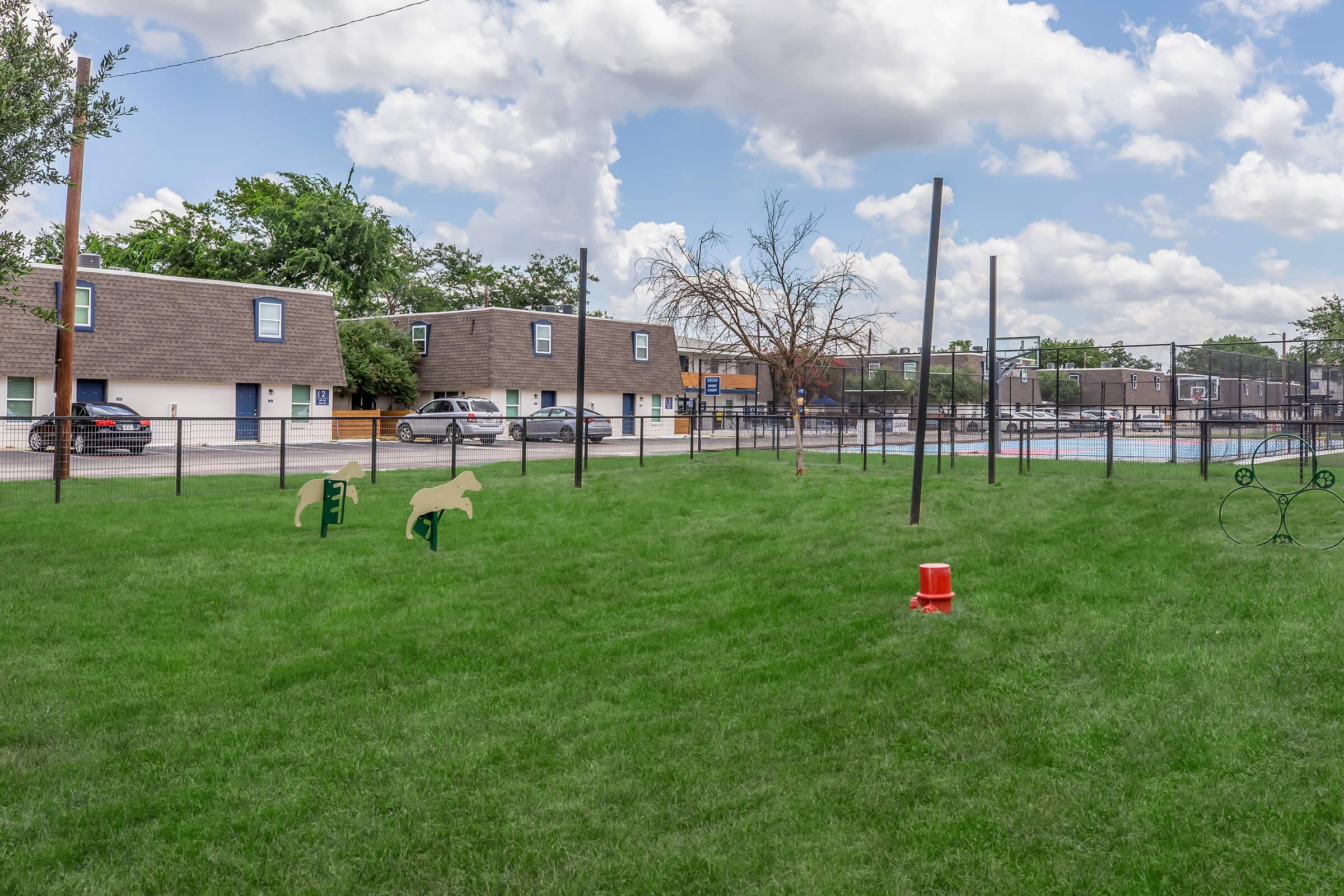 A grassy area with green playground equipment shaped like animals, including a horse and a dog. In the background, there are several buildings and a fenced tennis court. The sky is partly cloudy, adding to the bright atmosphere of the scene.