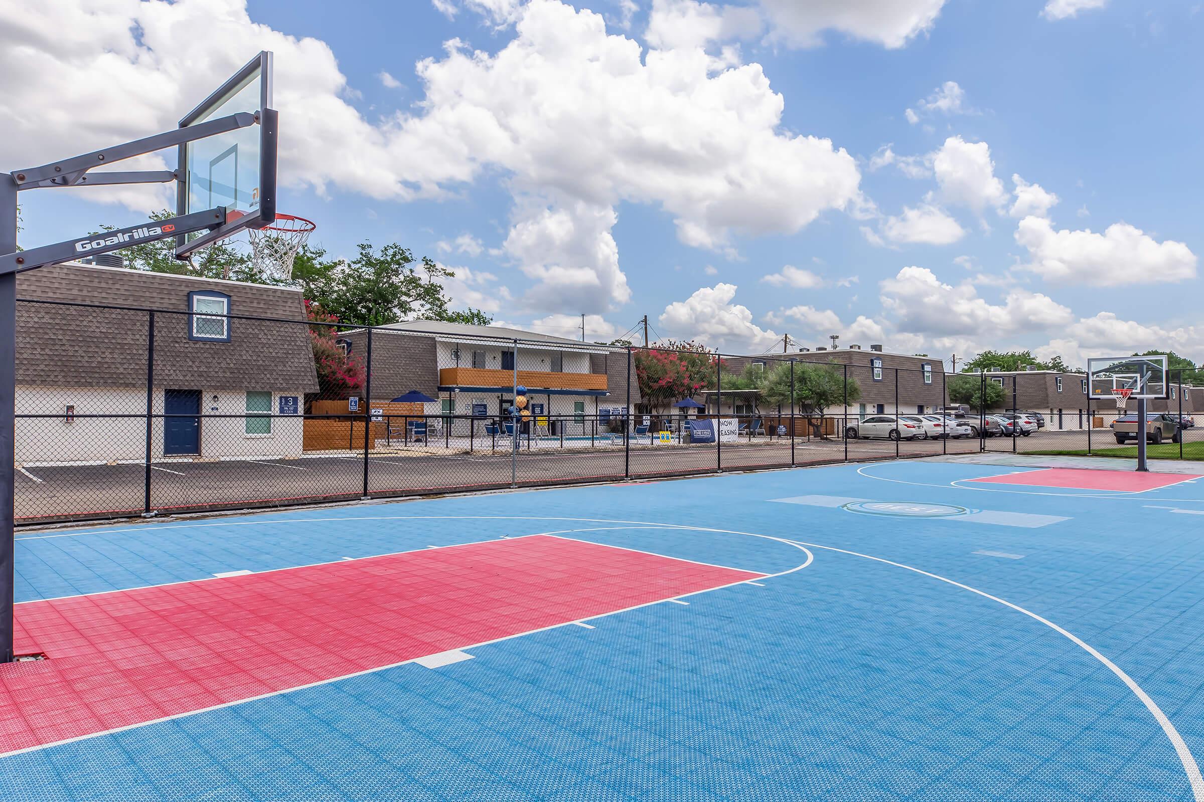 A colorful basketball court featuring red and blue playing surfaces, surrounded by a chain-link fence. In the background, there are several buildings and trees under a bright, partly cloudy sky. Two basketball hoops are visible at either end of the court.