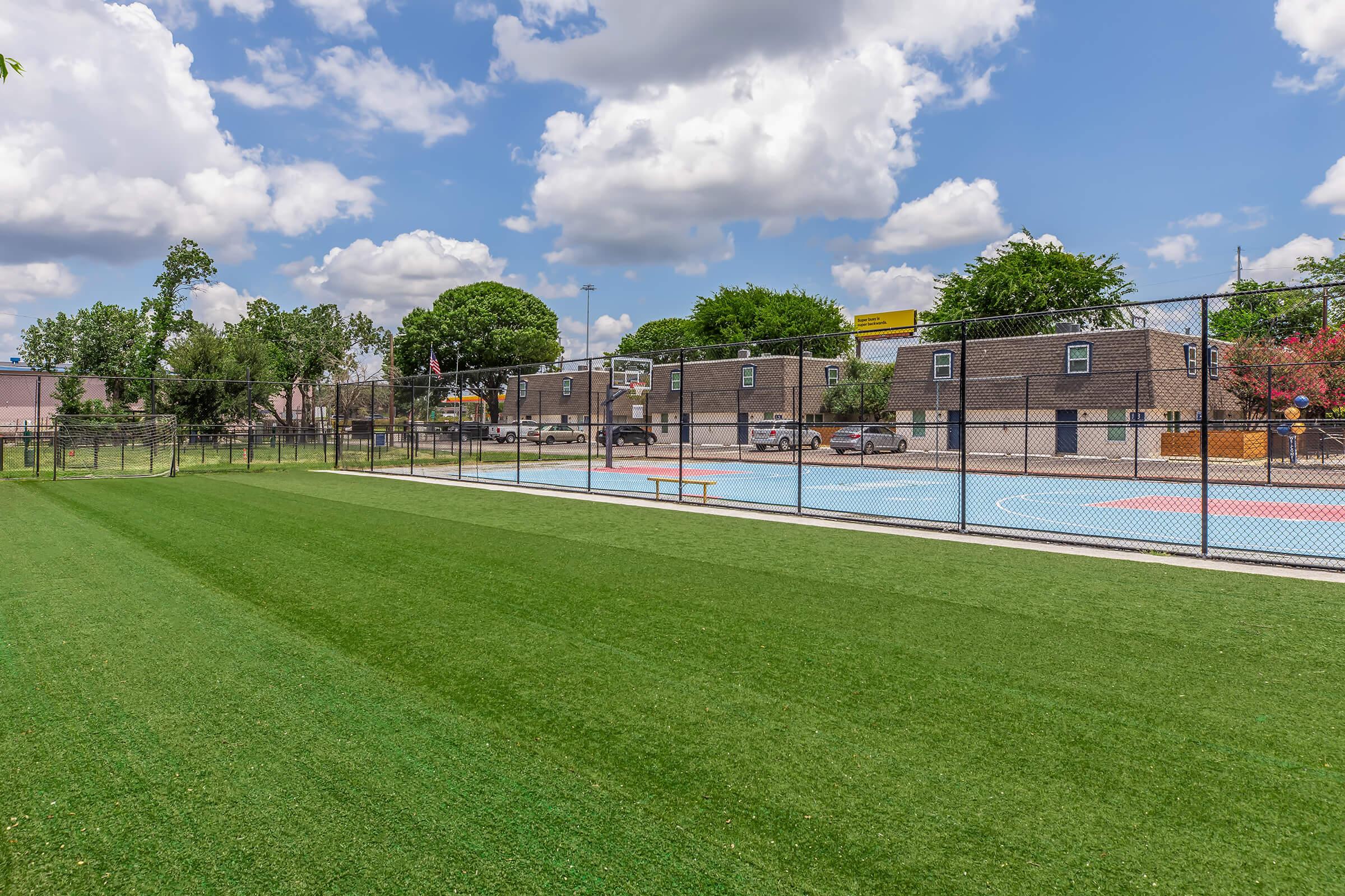 A landscaped sports area featuring a fenced basketball court and a colorful hardcourt, surrounded by greenery. The sky is mostly cloudy, and there are residential buildings in the background, creating a vibrant community atmosphere.