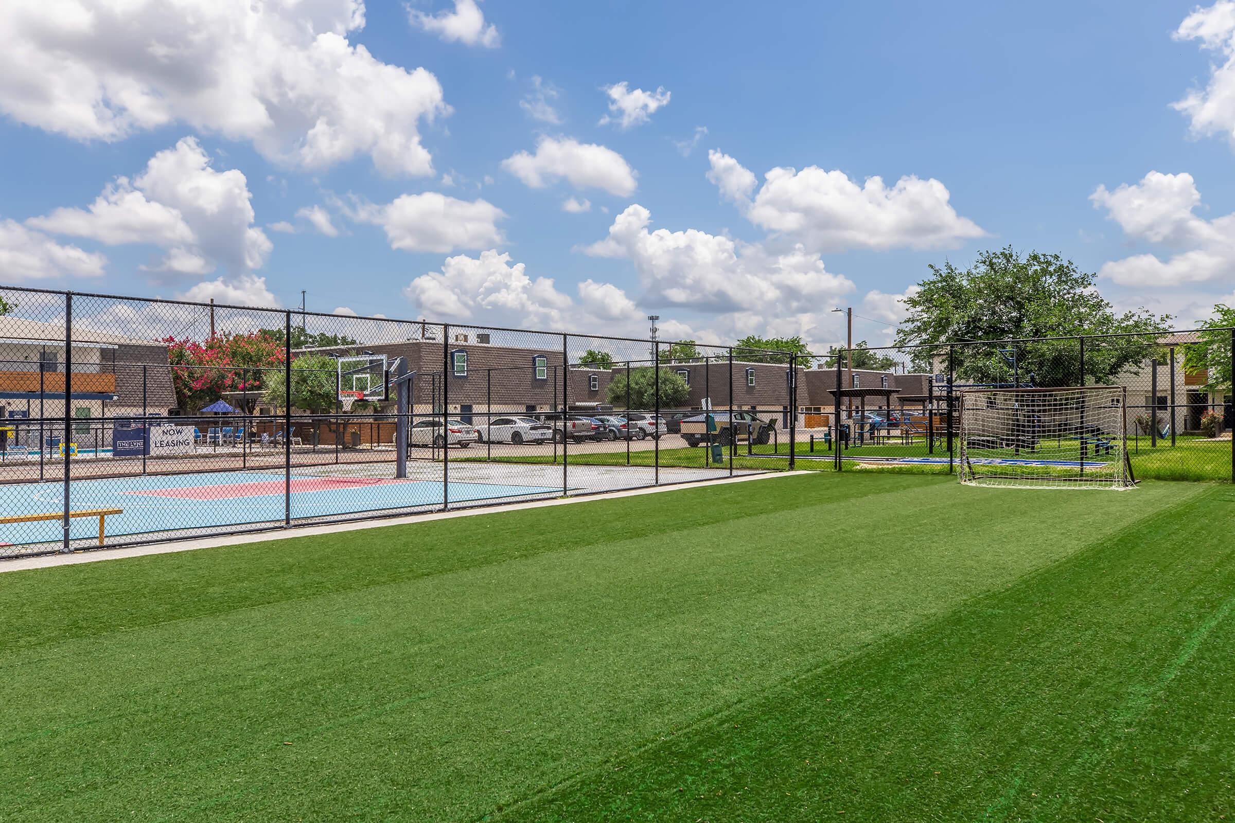 A view of a multi-sport facility featuring a basketball court and a soccer field. The basketball court is fenced and has colorful markings, while the soccer area has artificial grass. In the background, there are trees and buildings under a bright blue sky with fluffy white clouds.