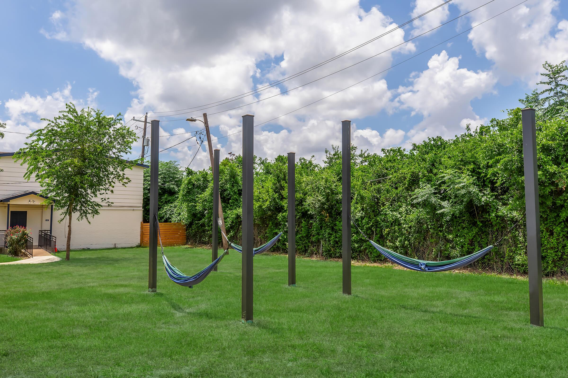 A tranquil outdoor space featuring several blue hammocks suspended between tall posts, set against a backdrop of green foliage and a partly cloudy sky. The grassy area invites relaxation and enjoyment of nature, with utility poles adding a functional element to the scene.