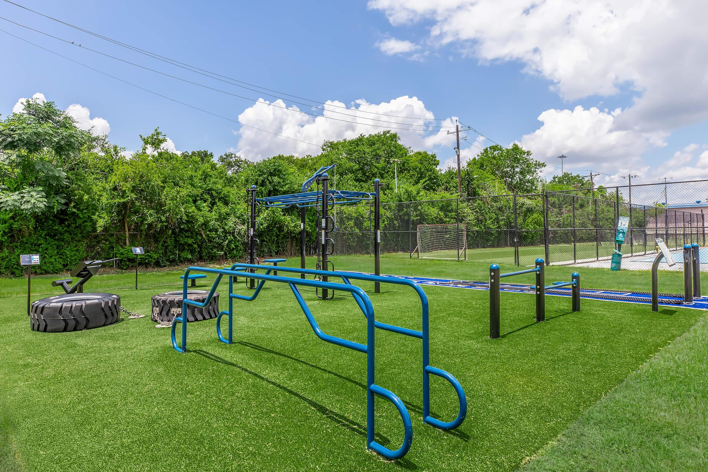 Outdoor fitness area featuring blue workout equipment on artificial turf. Includes climbing bars, a tire for flipping, and various exercise stations. Behind the area, there are trees and a soccer field visible in a sunny sky with a few clouds.