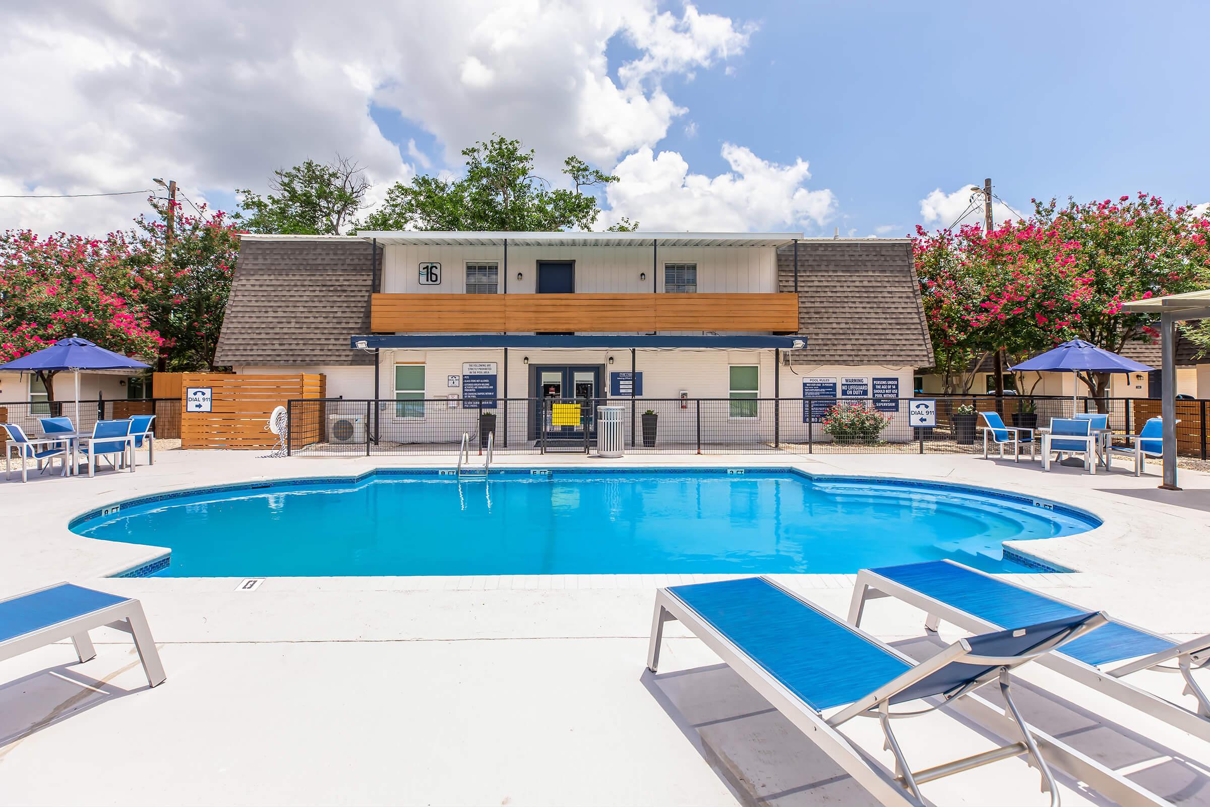 A sparkling blue pool surrounded by lounge chairs, with a building in the background featuring a wooden facade and vibrant greenery. The scene includes umbrellas for shade, and a clear sky with scattered clouds, creating a relaxing outdoor atmosphere.