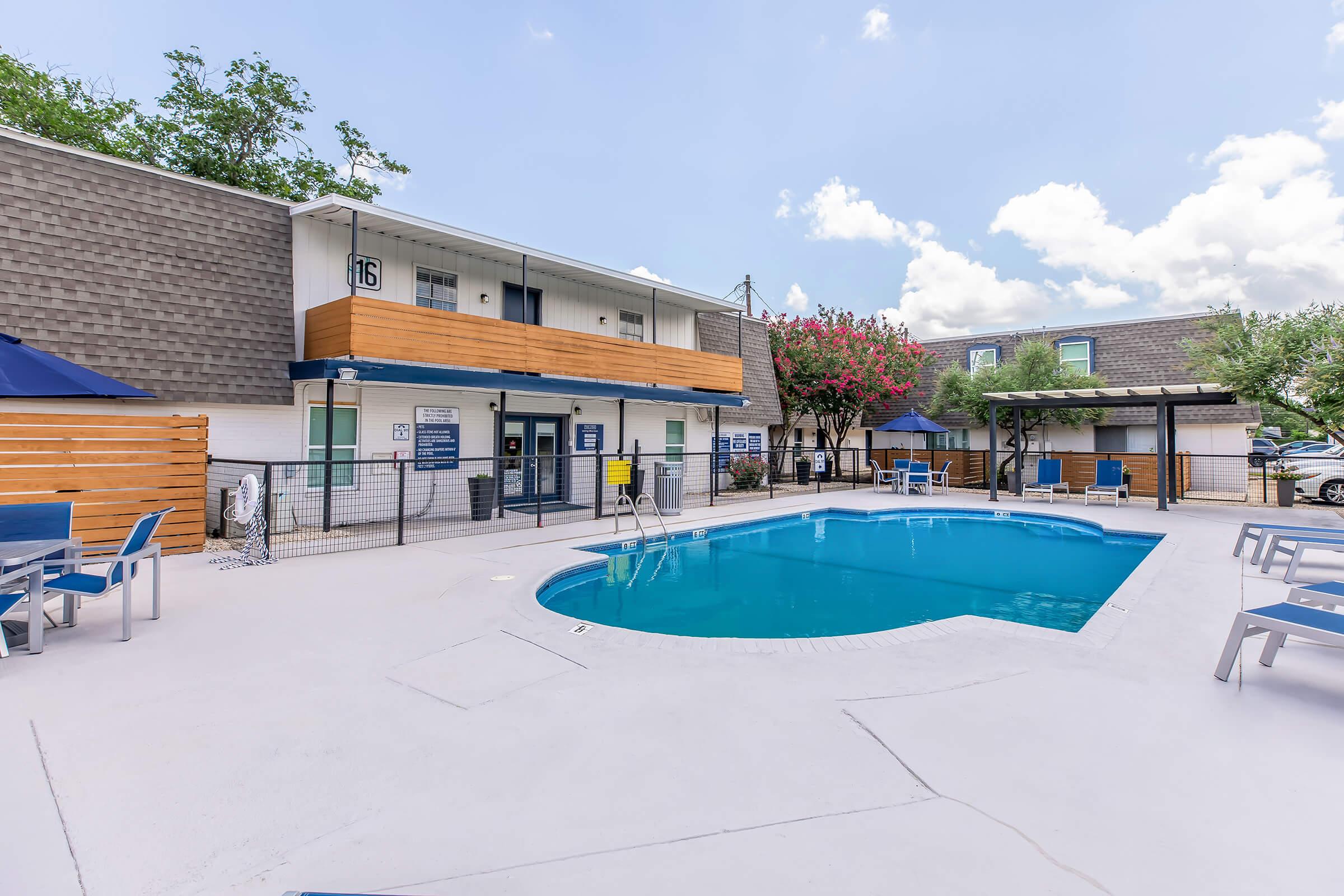 A picturesque hotel courtyard featuring a sparkling blue pool surrounded by lounge chairs. In the background, there are two levels of accommodations, with wooden balconies and vibrant trees nearby. The scene is bright and inviting under a partly cloudy sky.
