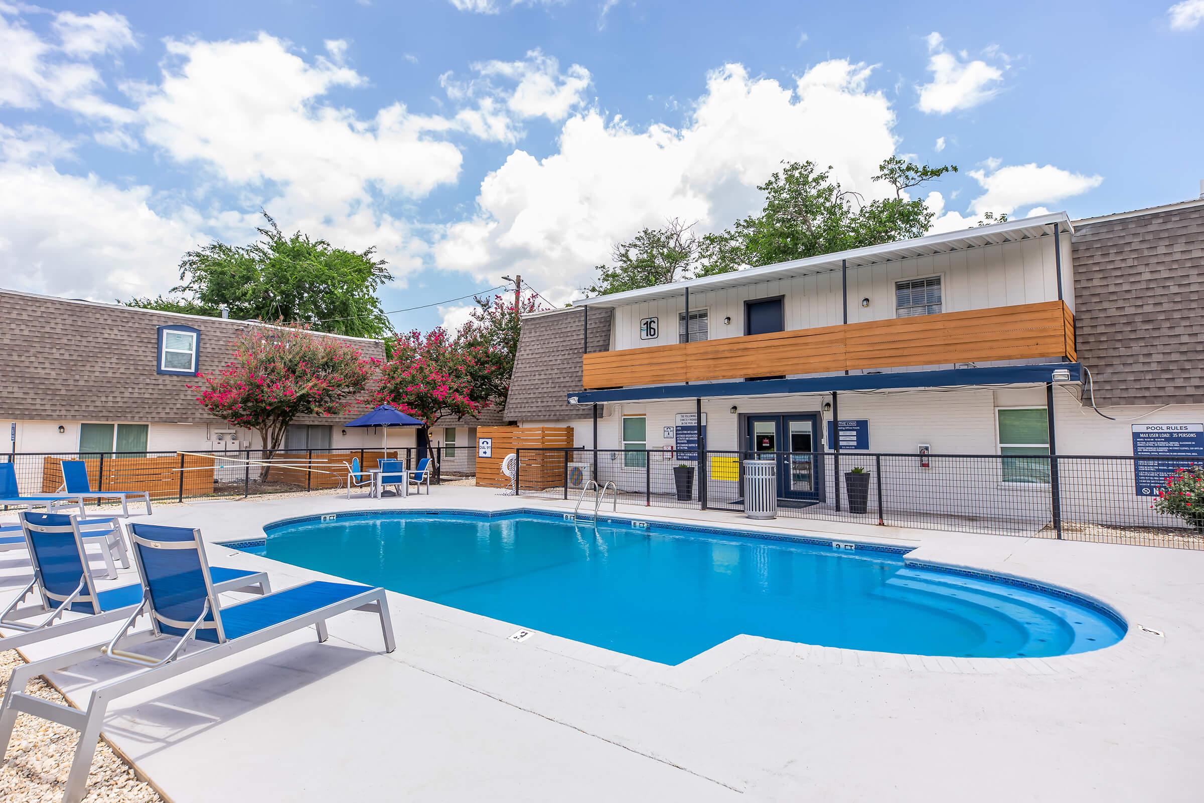 A clear view of a swimming pool surrounded by lounge chairs, near a well-maintained building with a balcony. The area is adorned with greenery and features partly cloudy blue skies, creating a relaxing and inviting atmosphere.