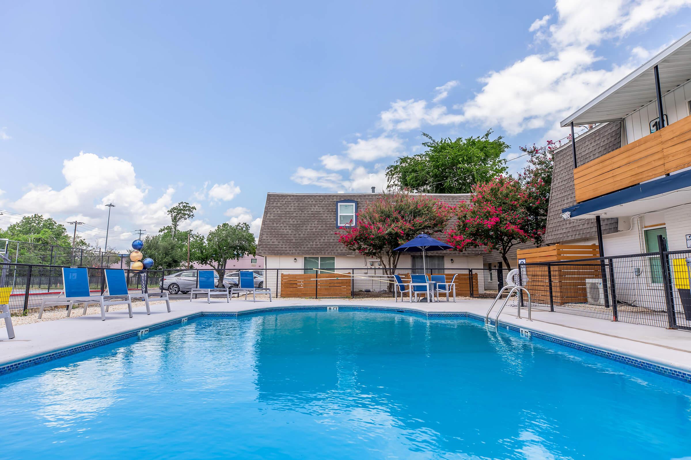 A clear blue swimming pool surrounded by lounge chairs under a sunny sky. In the background, there is a multi-story building with wooden accents and flowering trees. The atmosphere is inviting and relaxing.