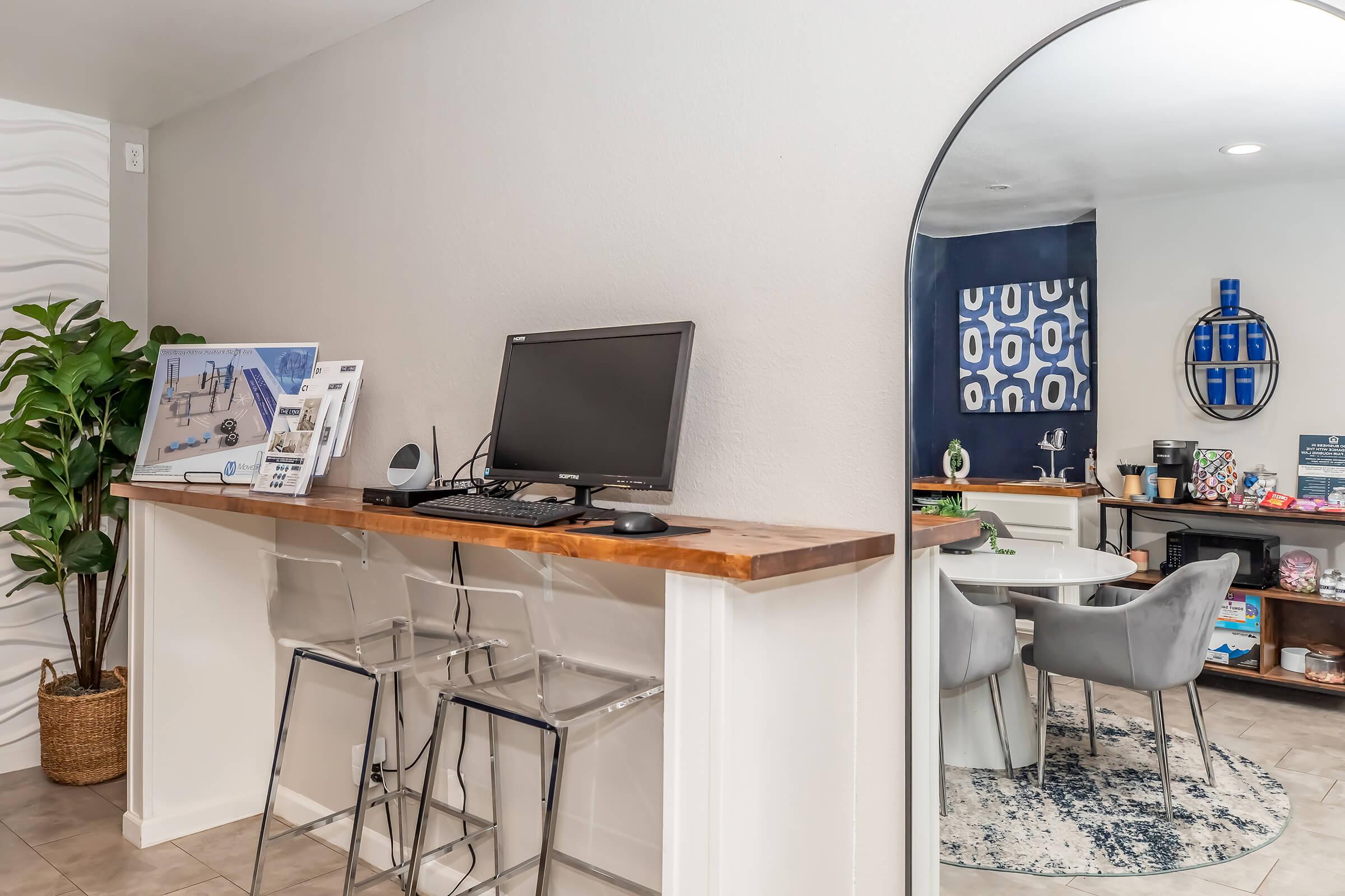 A modern workspace featuring a clear bar with two acrylic stools, a computer monitor, and a plant. In the background, there's a cozy dining area with a round table and chairs, a stylish rug, and wall decorations, creating a welcoming atmosphere.