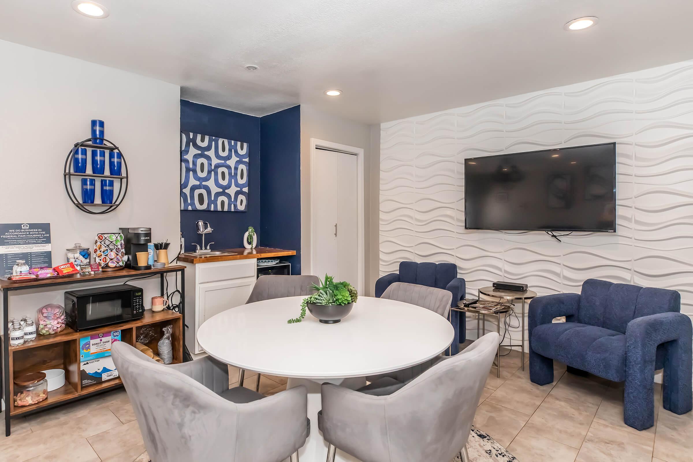 Modern living area featuring a round white dining table with gray upholstered chairs. A blue accent wall with geometric patterns is behind a large flat-screen TV mounted on a textured white wall. A small kitchenette and a cart with kitchen appliances and snacks are visible, enhancing the cozy atmosphere.