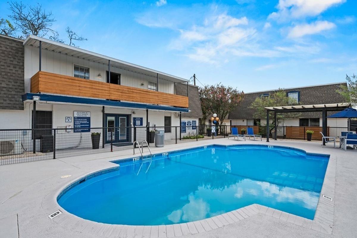 A clean outdoor swimming pool surrounded by a fence, with lounge chairs and tables nearby. There are two buildings in the background, one with a wooden balcony. The sky is clear with a few clouds, creating a bright and inviting atmosphere.