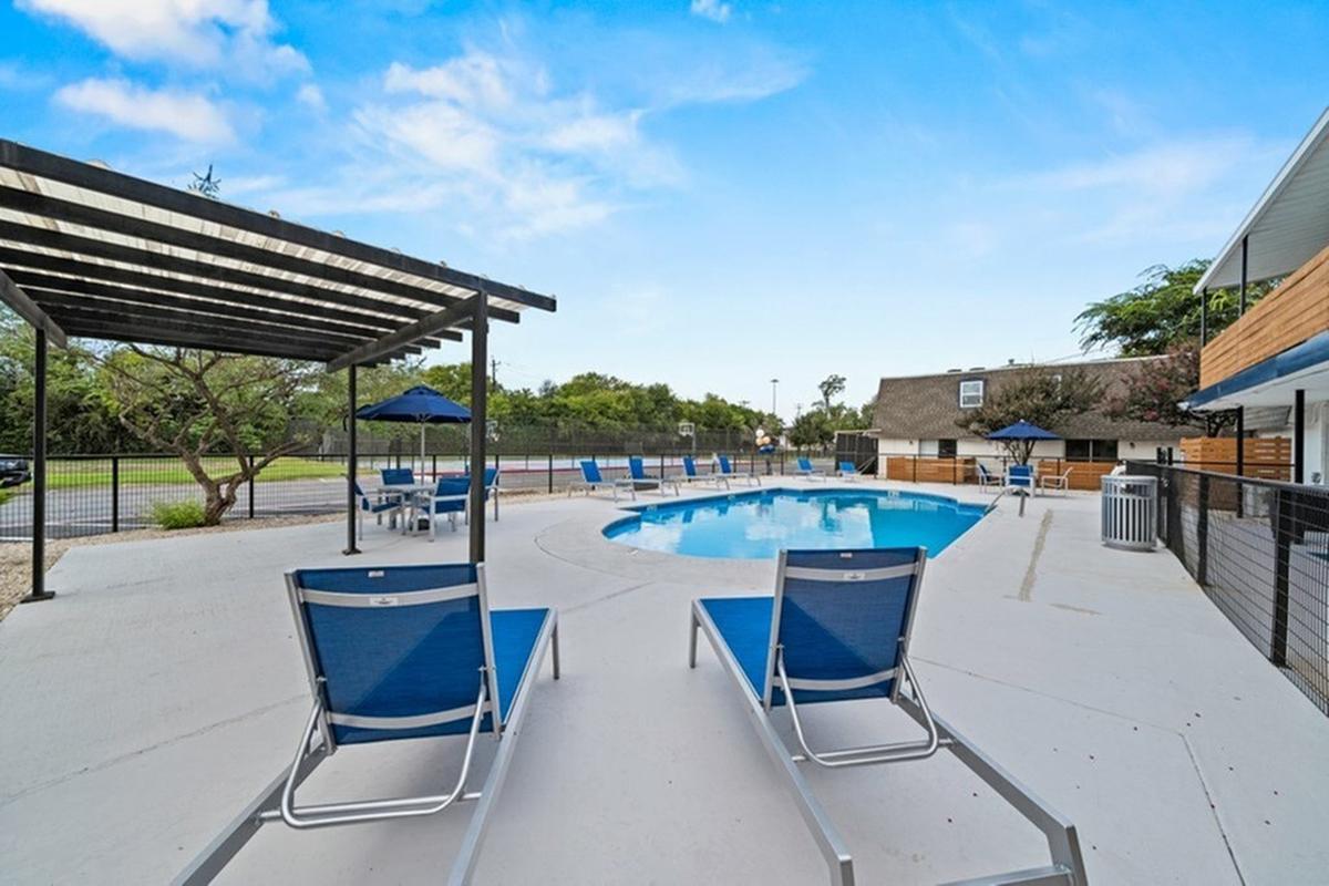 Bright, inviting pool area with two blue lounge chairs placed near the edge of a crystal-clear swimming pool. The pool is surrounded by a fence, with a pergola overhead and blue umbrellas providing shade. Lush greenery is visible in the background under a clear blue sky.