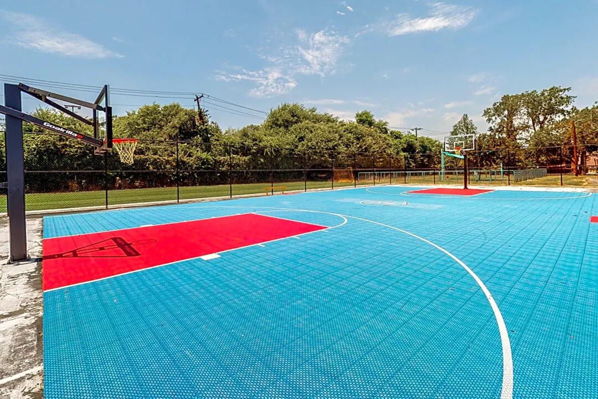 A brightly colored outdoor basketball court featuring blue and red surfaces, with a basketball hoop on one end. The court is surrounded by a fence and lush greenery in the background under a clear blue sky.