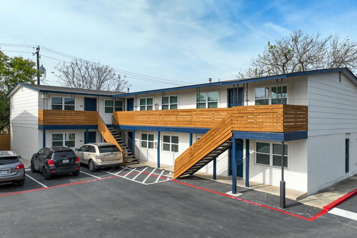 Two-story building with a modern exterior featuring wooden stairs and railings. Ground-level parking spaces are visible in front, with several cars parked. The building has large windows and a clean, well-maintained look, set against a clear blue sky. Trees are partially visible in the background.