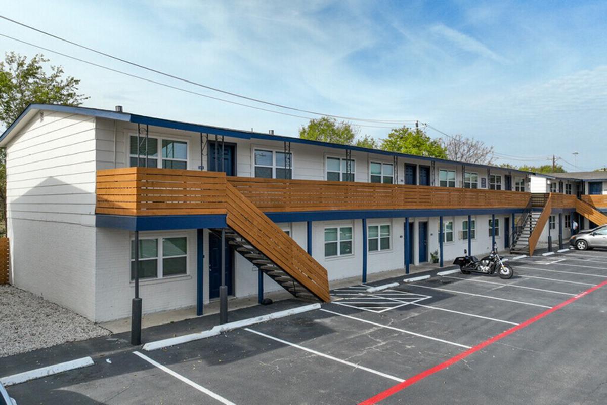 A low-rise motel building featuring two levels with wooden staircases, surrounded by a paved parking lot. The building has a modern design with a blue and white color scheme, and several windows. A motorcycle is parked nearby, and the landscape includes a few trees. The sky is clear and blue.