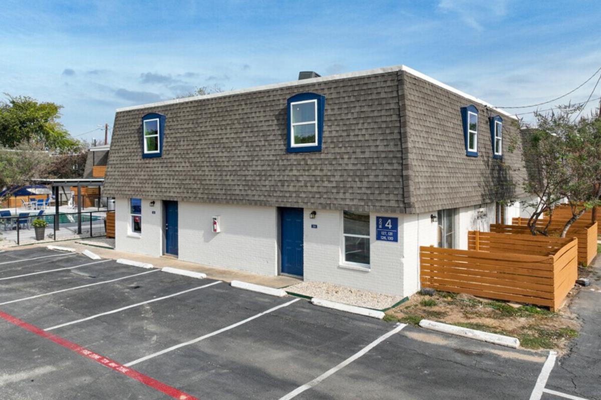 A two-story building with a sloped roof, featuring light-colored walls and dark blue window frames. The exterior is clean with a parking lot in front, marked spaces, and a small wood fence to one side. There are trees in the background and a play area visible in the distance.