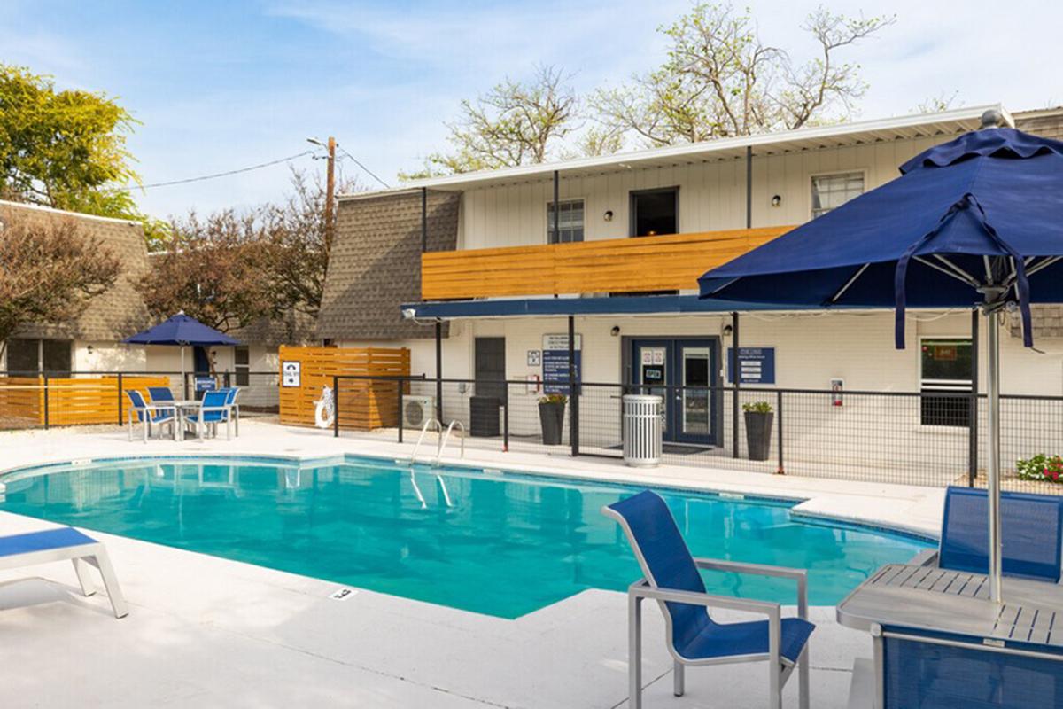 A clear blue swimming pool surrounded by lounge chairs and umbrellas. In the background, there is a two-story building with a wooden balcony. The area is landscaped with trees and greenery, creating a relaxing ambiance. The scene is well-lit, indicating a sunny day.
