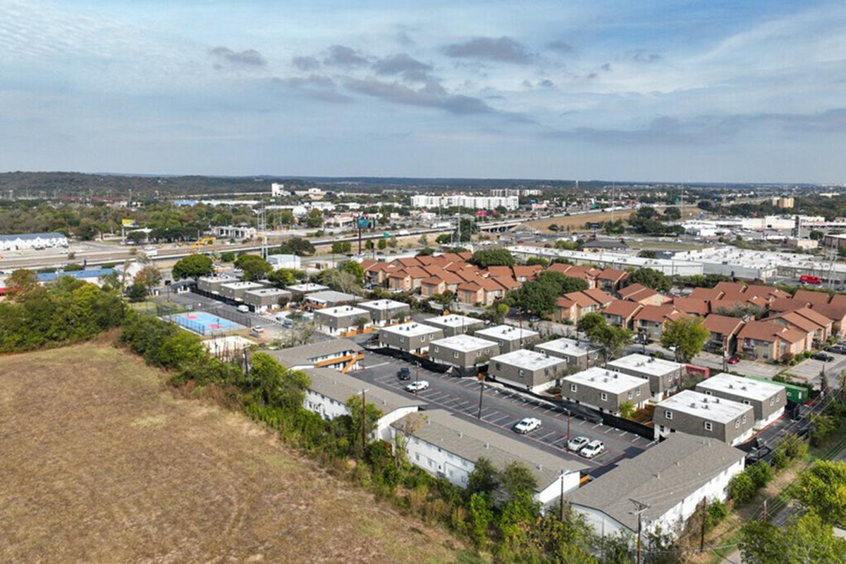 Aerial view of a suburban area featuring a mix of residential complexes and commercial buildings. The landscape includes a parking lot, grassy fields, and distant highway infrastructure, with trees lining the edges of the developed area under a partly cloudy sky.