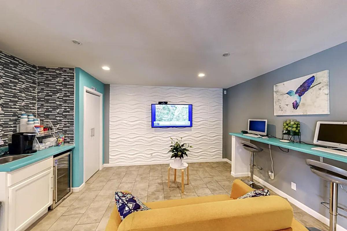 A modern lounge area featuring a light-colored wall with wave patterns, a large flat-screen TV mounted on the wall, two computers on a shelf, and a small table with a flower arrangement. The space includes a yellow couch with colorful cushions and a kitchenette area with a water cooler.