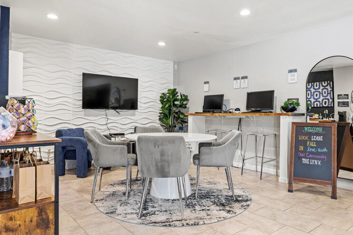Modern office space featuring a round white table surrounded by gray chairs on a patterned rug. A large TV is mounted on a textured wall, with a computer station in the background. Decorative plants add a touch of greenery, while a welcome sign on a nearby board greets visitors.