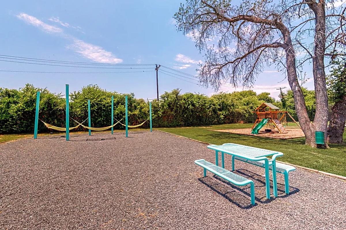 A sunny playground featuring a slide and climbing structure on one side, and a gravel area with yellow swings on the other. There are two turquoise benches nearby, surrounded by green grass and trees in the background, creating a cheerful outdoor environment.