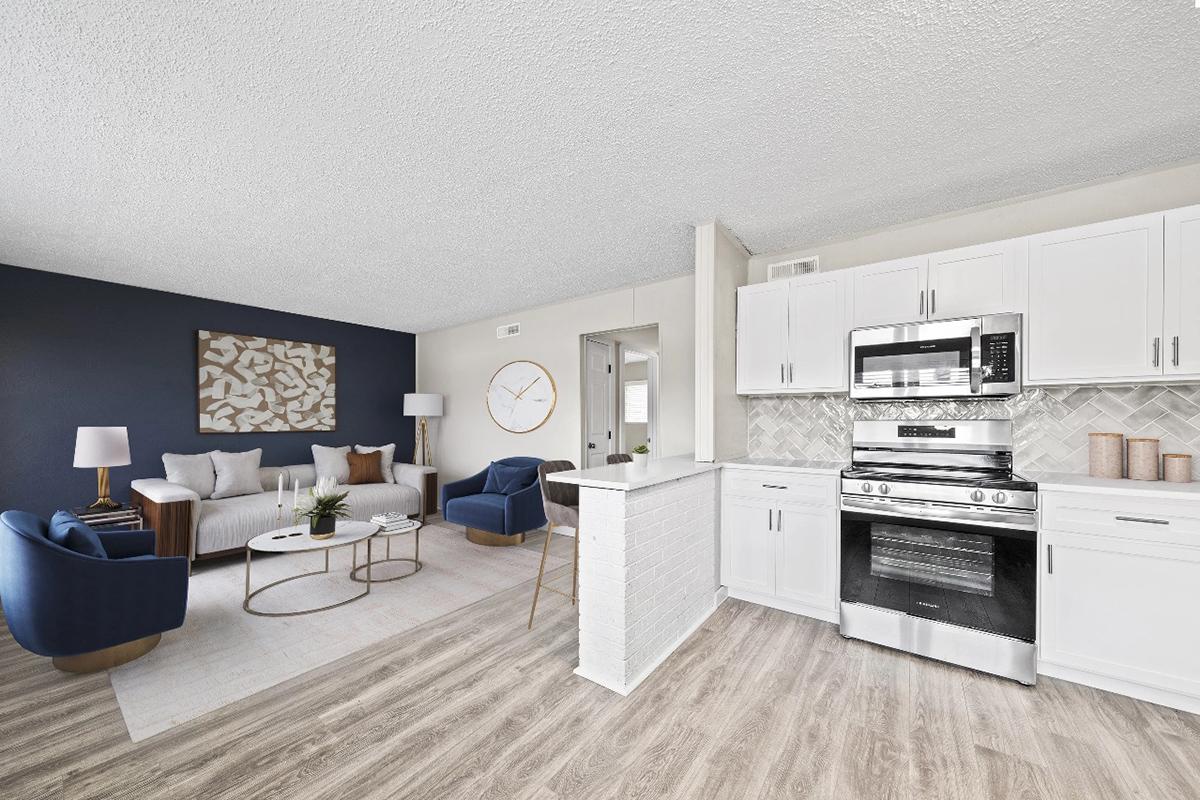 Modern living room and kitchen area featuring a cozy seating arrangement with blue chairs and a white sofa. A stylish coffee table sits in the center, while a sleek stove and cabinetry are visible in the kitchen space. Light-colored walls and wooden flooring create a bright, inviting atmosphere.