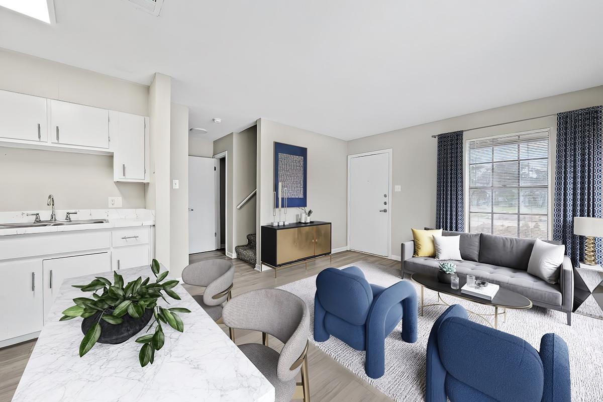 A modern living room and kitchen area featuring a marble-topped table, a gray sofa, blue accent chairs, and a decorative plant. Natural light fills the space through large windows, and a staircase is visible in the background, leading to a second floor. Stylish décor and neutral colors create a welcoming atmosphere.