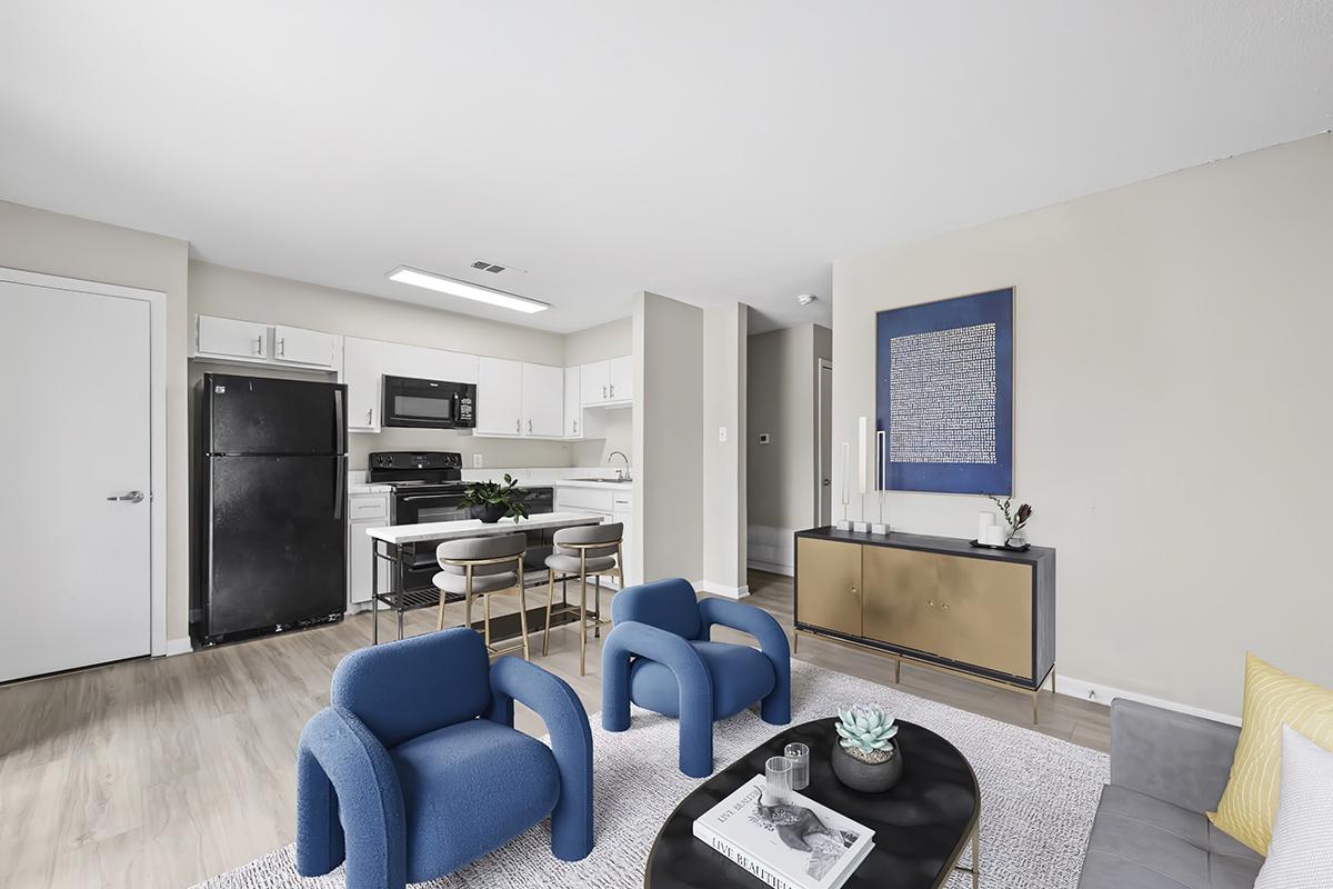 A modern living room and kitchen area featuring light-colored walls, a black refrigerator, and stainless steel appliances. The living space includes a gray rug, a low coffee table with decorative items, and two blue armchairs. A gold sideboard with a blue wall art piece completes the stylish decor.